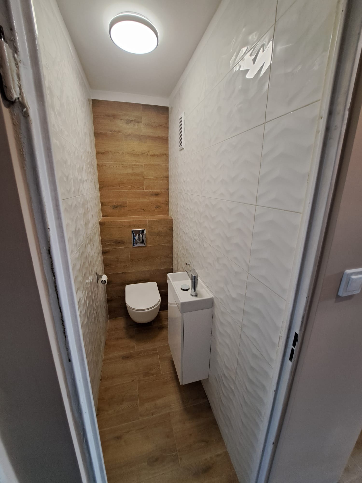 Mała łazienka po remoncie: białe płytki z falistym wzorem na ścianach, brązowe płytki imitujące drewno na podłodze i ścianie za toaletą, wisząca toaleta i umywalka z szafką, widok zza otwartych...