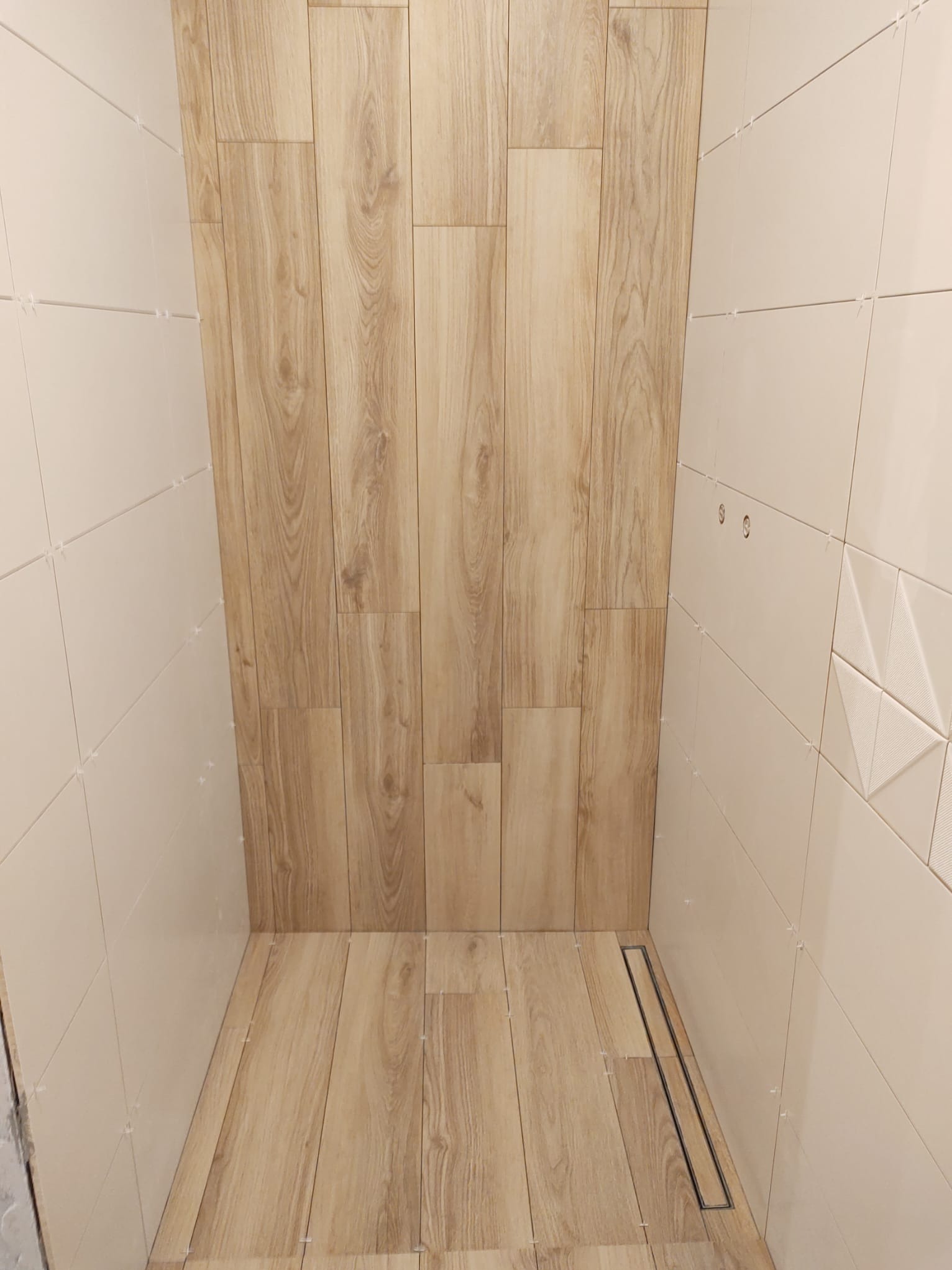 Wnętrze kabiny prysznicowej w trakcie wykańczania, z drewnopodobnymi płytkami na ścianach i podłodze, jasne płytki na pozostałych ścianach z dekoracyjnym wzorem, widoczne krzyżyki dystansowe...