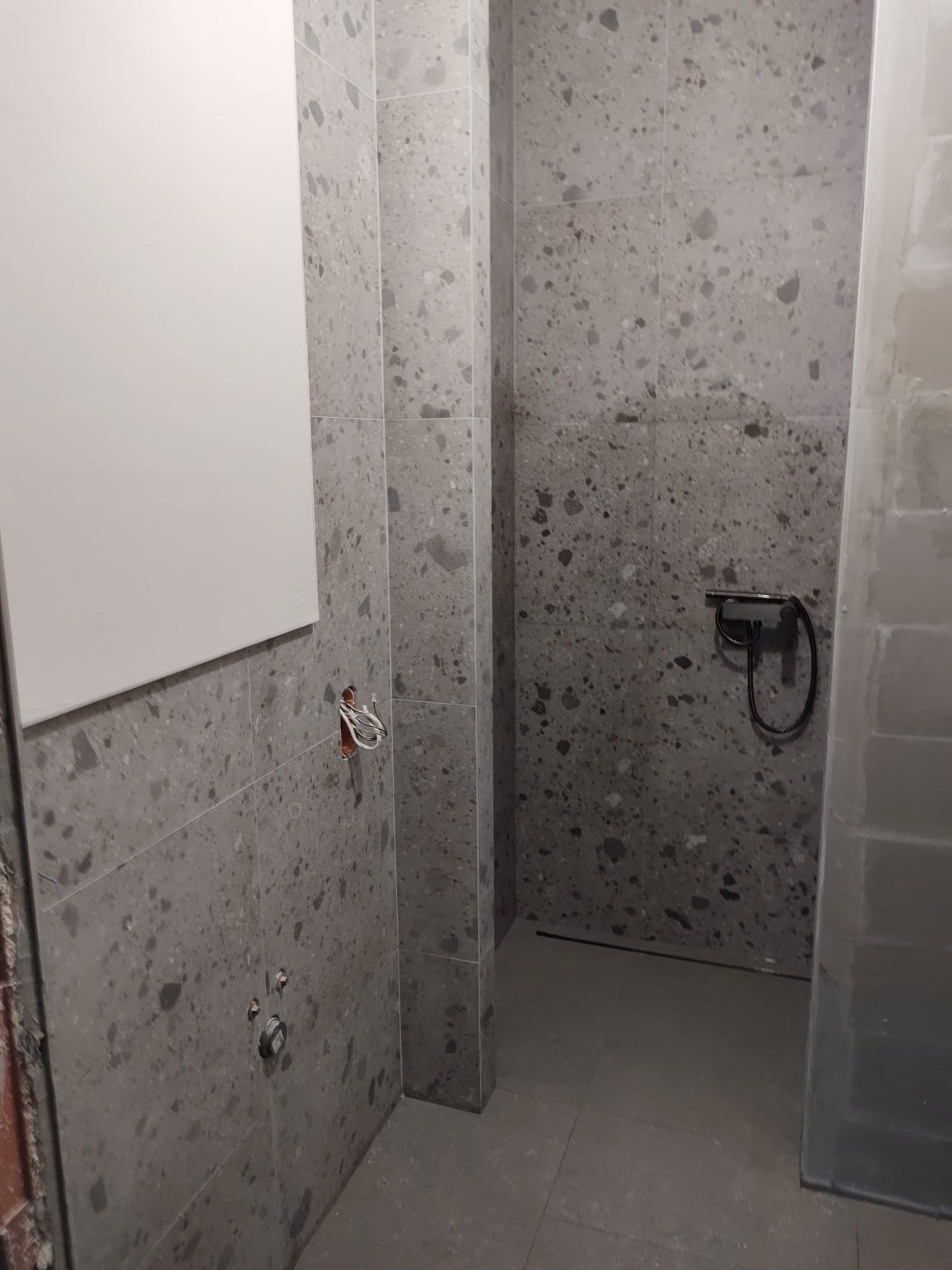 Wnętrze łazienki w trakcie wykańczania, ściany i podłoga wyłożone szarymi płytkami imitującymi lastryko, widoczne niedokończone instalacje hydrauliczne i elektryczne, oraz biały grzejnik na ścianie.