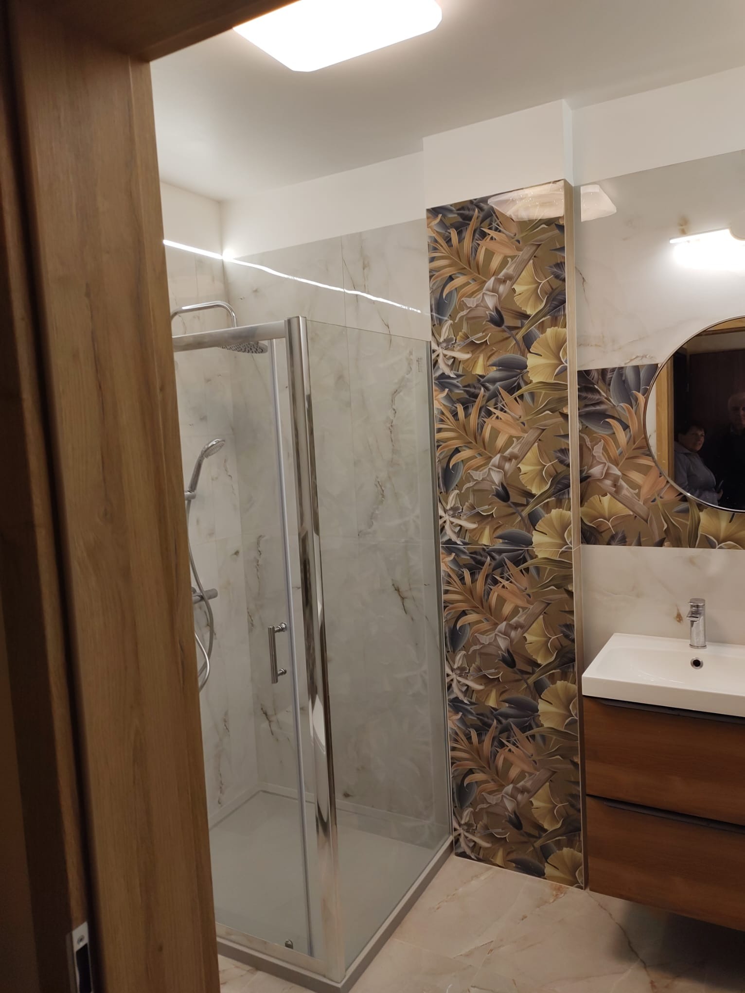 Nowoczesna łazienka z kabiną prysznicową, umywalką z szafką z drewna oraz płytkami z motywem roślinnym na ścianie, widok zza drewnianej ościeżnicy drzwi.