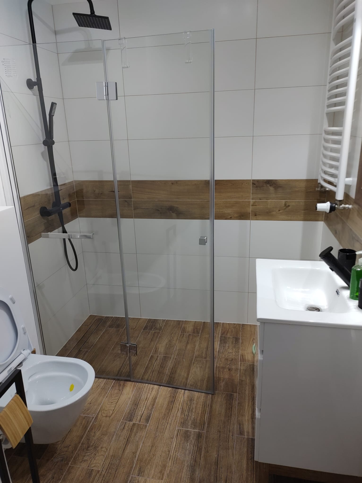 Nowoczesna łazienka z kabiną prysznicową typu walk-in, czarną armaturą i płytkami imitującymi drewno na podłodze oraz w pasie na ścianach, widoczna umywalka z szafką i toaleta z deską w kolorze...