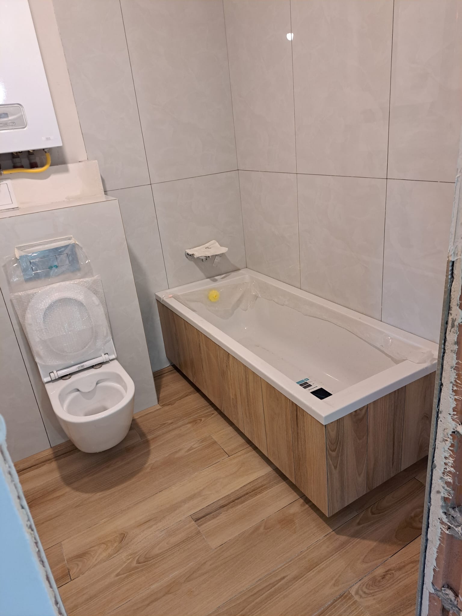 Wnętrze łazienki w trakcie wykańczania: biała wanna obudowana panelami imitującymi drewno, toaleta jeszcze w folii ochronnej, jasne płytki na ścianach i podłodze, widoczny piec gazowy.