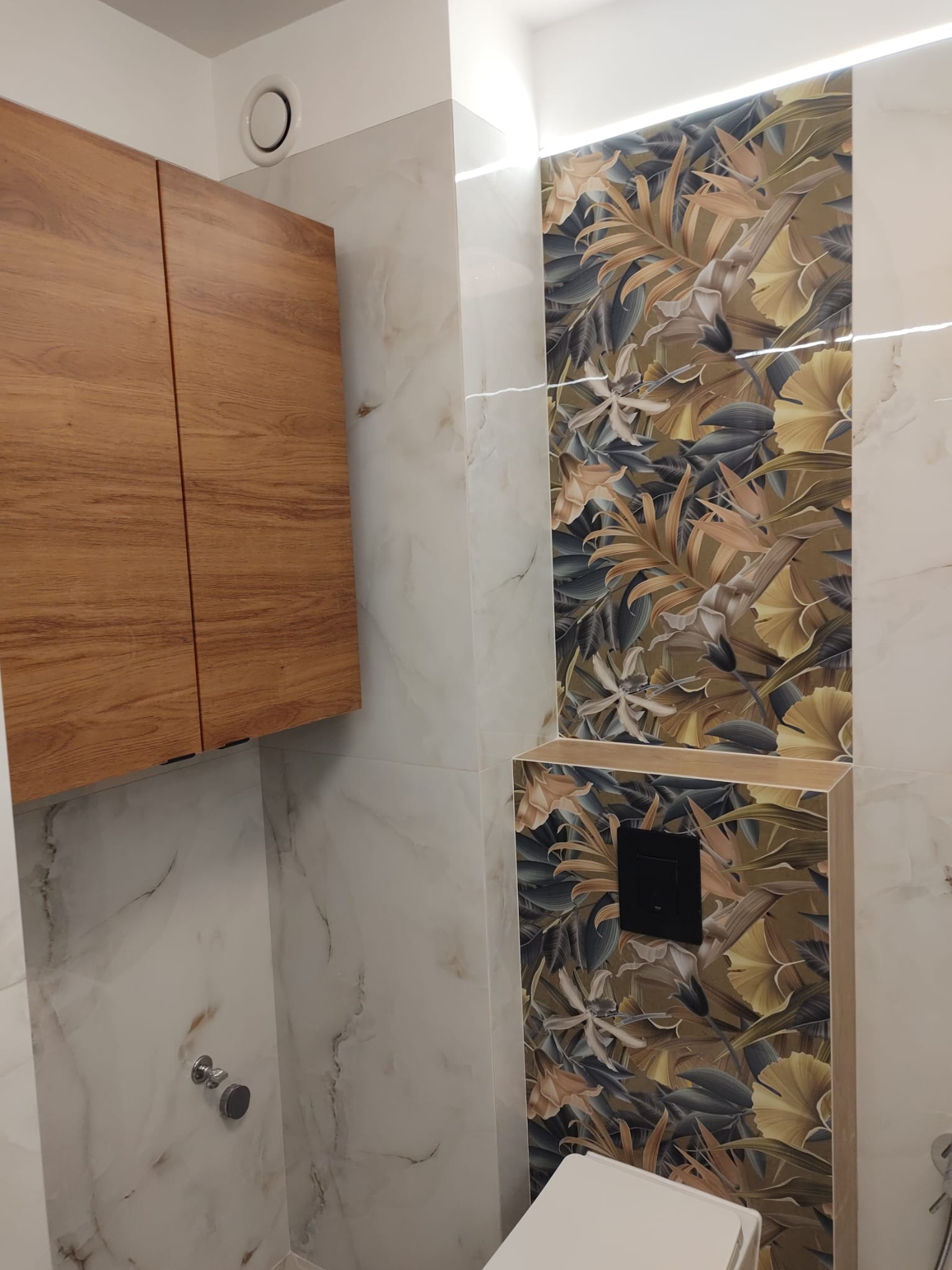 Nowoczesna łazienka z drewnianą szafką wiszącą, marmurowymi płytkami ściennymi i panelami z motywem tropikalnych liści, oświetlona subtelnym światłem.