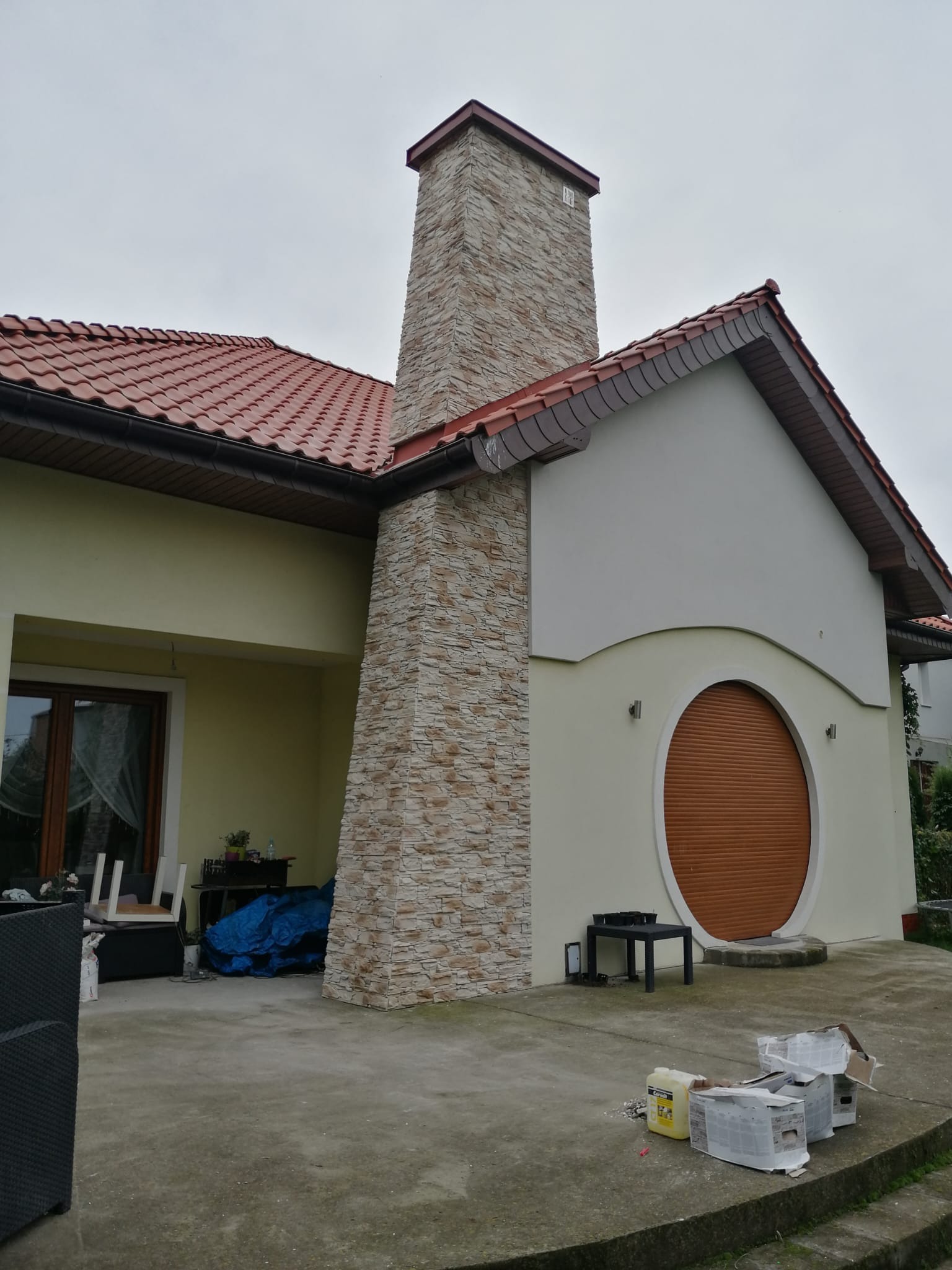 Elewacja domu jednorodzinnego z kominem wykończonym kamieniem dekoracyjnym, widoczne puszki i kartony po materiałach budowlanych na tarasie.