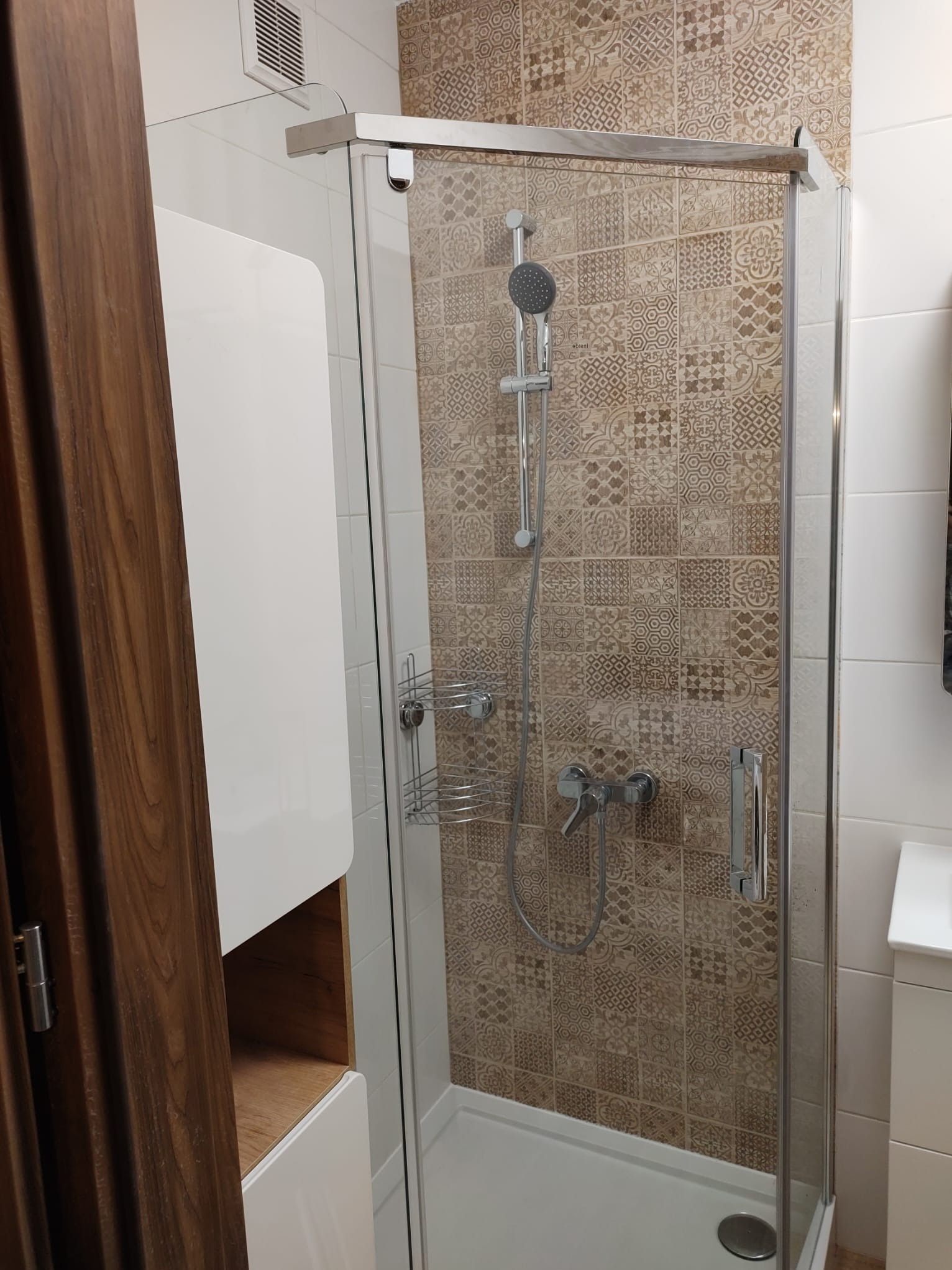 Kabina prysznicowa z deszczownicą i wzorzystymi brązowymi płytkami w łazience w Piasecznie, widoczna również biała szafka i fragment drewnianych drzwi.