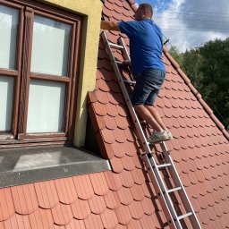 WEAVER STORK SPÓŁKA Z OGRANICZONĄ ODPOWIEDZIALNOŚCIĄ - Mężczyzna na drabinie przy oknie na dachu z czerwonej dachówki podczas prac na wysokości.