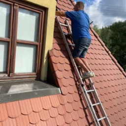 Mężczyzna na drabinie przy oknie na dachu z czerwonej dachówki podczas prac na wysokości.