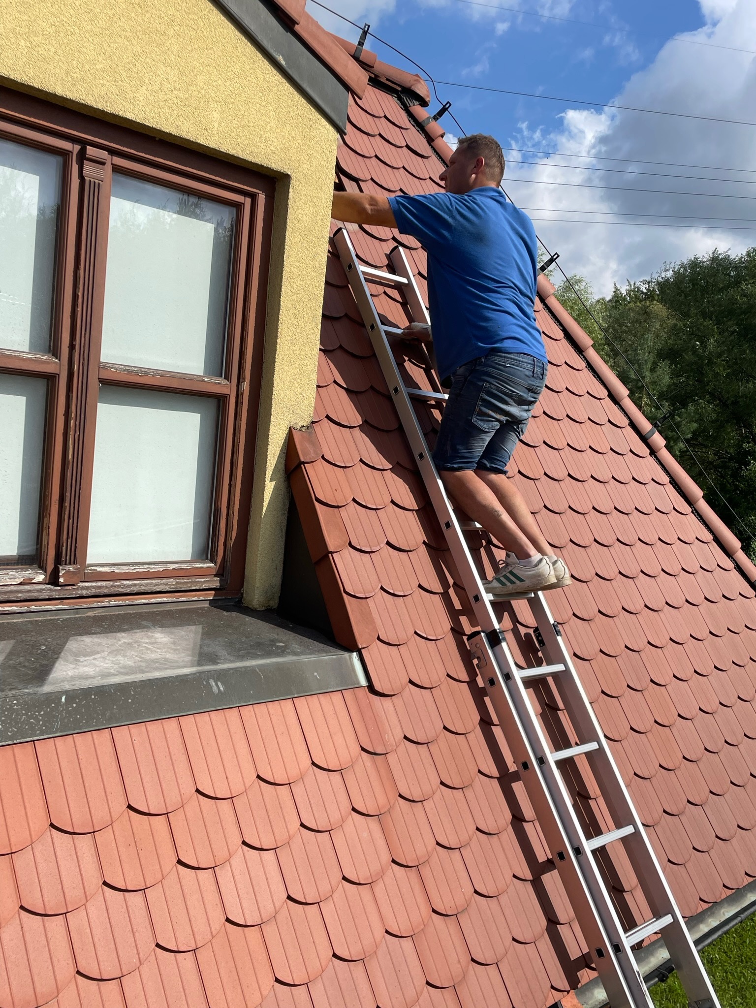 Mężczyzna na drabinie przy oknie na dachu z czerwonej dachówki podczas prac na wysokości.