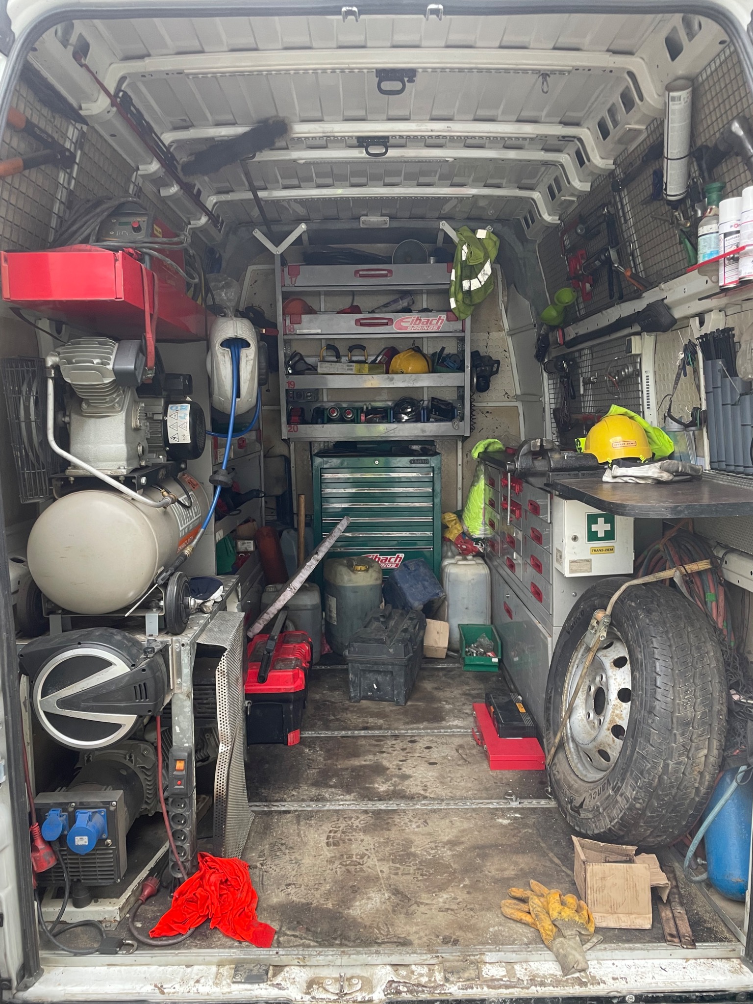 Wnętrze furgonetki serwisowej wypełnionej narzędziami, sprzętem i akcesoriami, w tym kompresorem, generatorem, skrzynkami narzędziowymi, kołem zapasowym i kaskiem ochronnym, z widocznymi śladami...