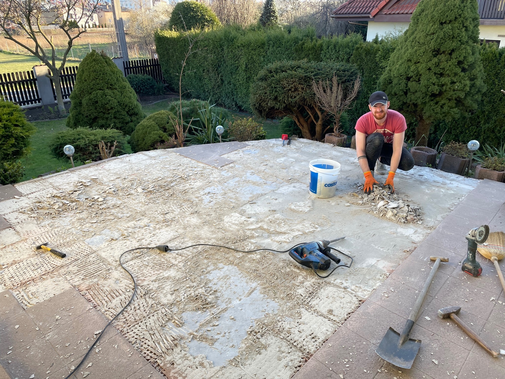 Mężczyzna w pomarańczowych rękawicach skuwa płytki z tarasu przy użyciu młotka udarowego, widoczne narzędzia: szpachelka, młotek, szpadel, szczotka, wiadro z zaprawą, w tle zadbany ogród z tujami.