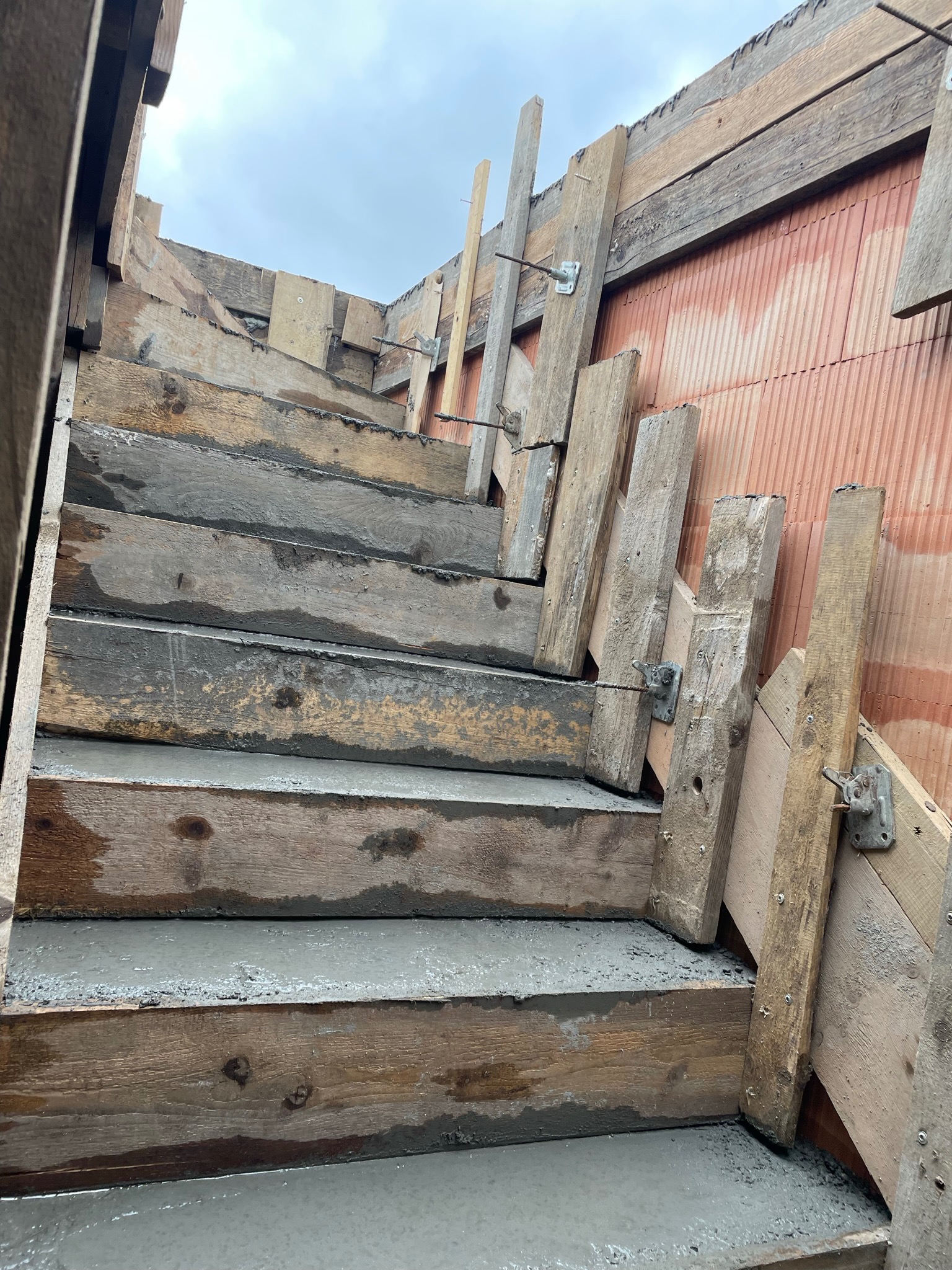 Betonowe schody w trakcie budowy, szalunek z desek, widoczne metalowe elementy mocujące, ściana z czerwonej cegły w tle.