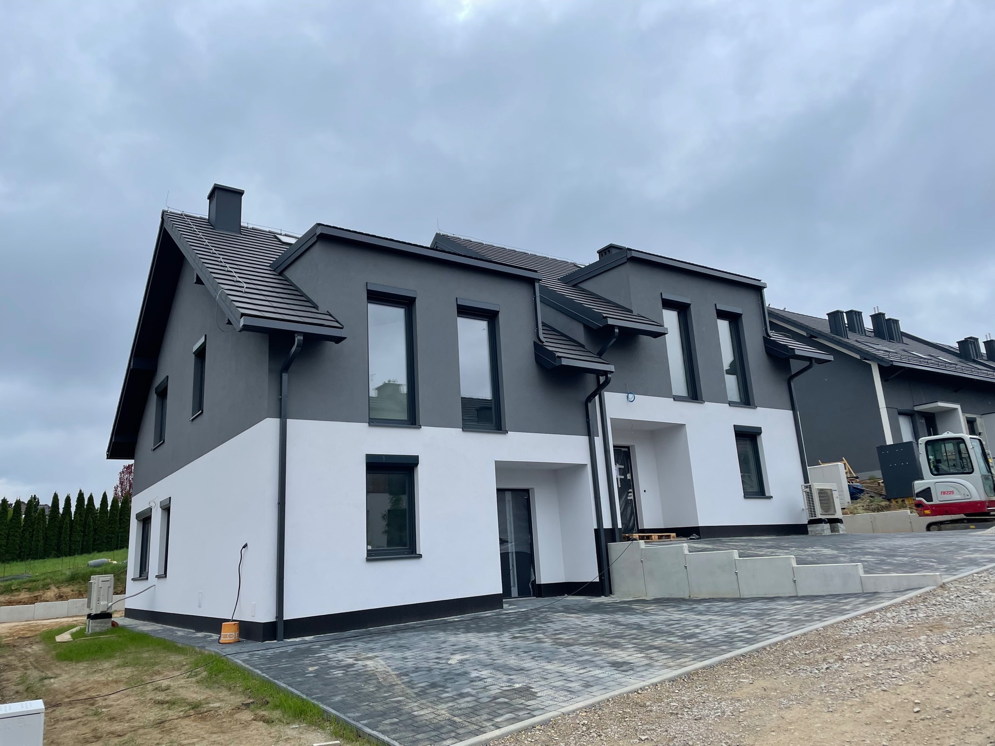 Nowoczesne szeregówki w dwukolorowej elewacji (szary i biały) z ciemną stolarką okienną i brukowaną nawierzchnią podjazdu, mała koparka widoczna w tle.