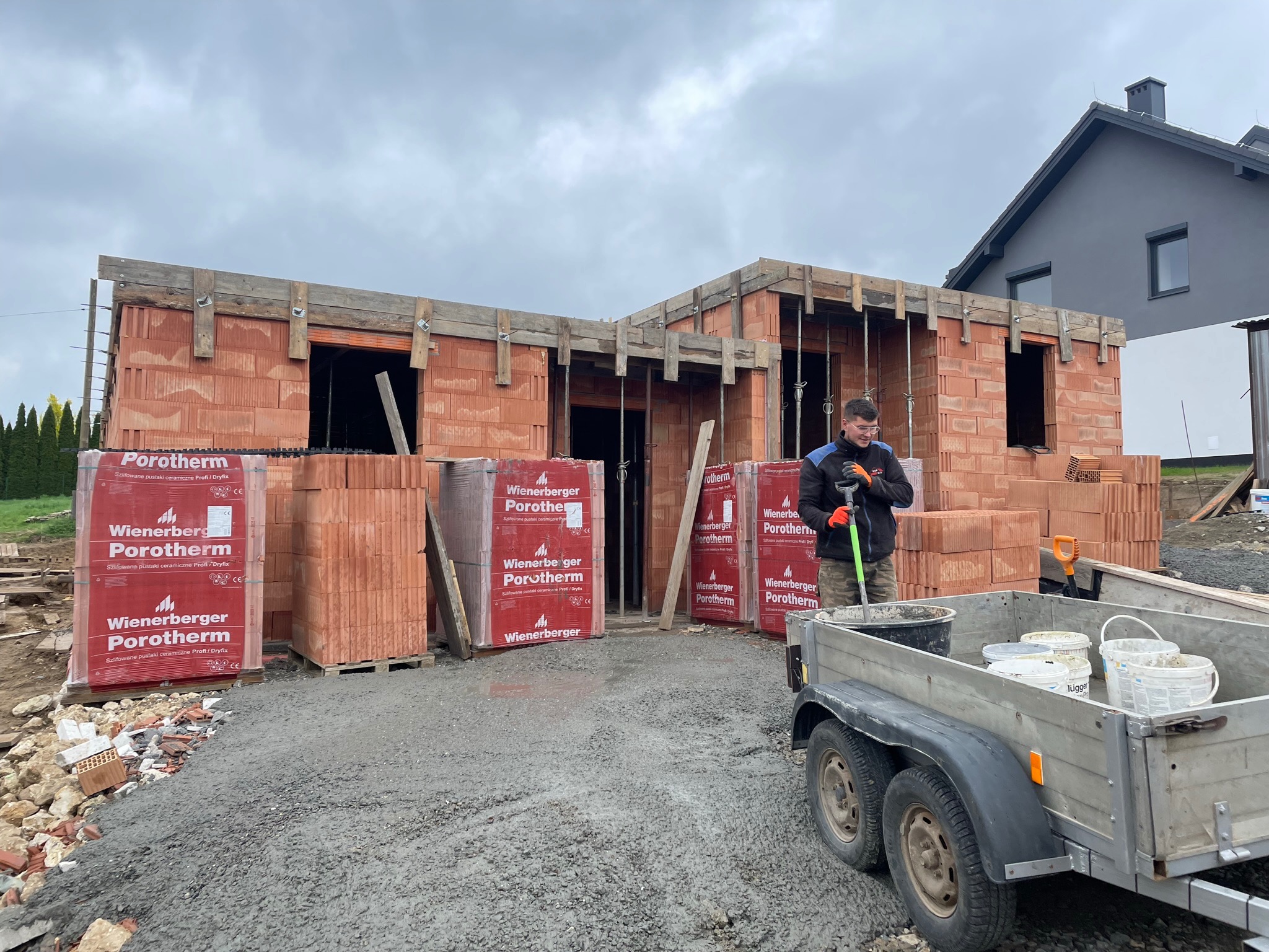 Wznoszenie murów z czerwonej cegły Porotherm na placu budowy, widoczne palety z cegłami, przyczepka z wiadrami i pracownik z łopatą na tle częściowo wybudowanego domu i sąsiedniego budynku.