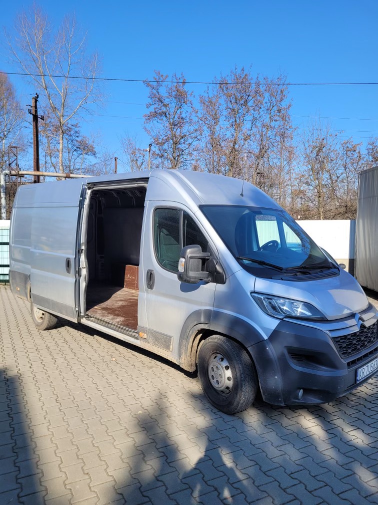 Srebrny bus dostawczy z otwartymi drzwiami bocznymi na kostce brukowej, widoczne drzewa bez liści w tle pod błękitnym niebem.