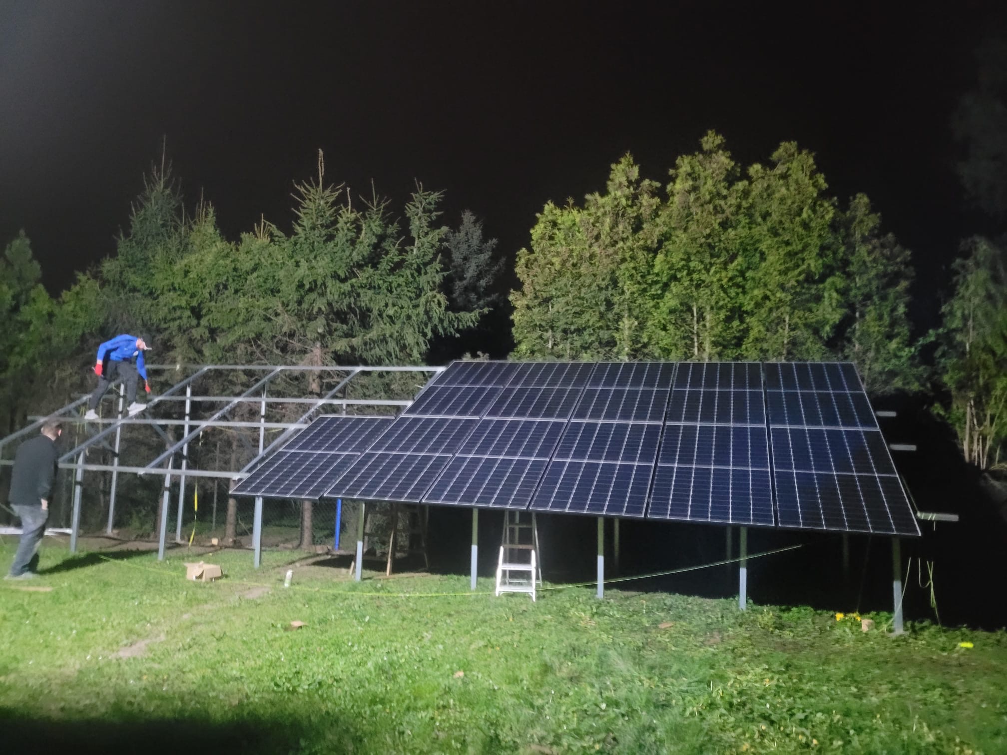 Montaż paneli fotowoltaicznych na metalowej konstrukcji w ogrodzie, widoczni dwaj pracownicy, jeden na rusztowaniu, drugi na trawniku, nocna sceneria.