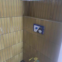 Marcin Kozaczuk - Pionowo ułożone beżowe płytki ścienne z ciemną fugą w narożniku łazienki, z widocznym systemem podtynkowym Geberit i elementami instalacji wodnej.