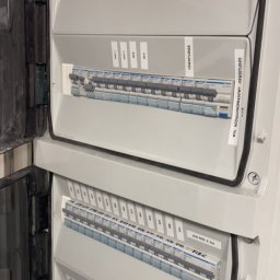ElektroNova Rafał Wnęk - Otwarta skrzynka elektryczna z bezpiecznikami i naklejkami opisującymi obwody, widoczne wyłączniki główne, UPS i obwody oświetleniowe.