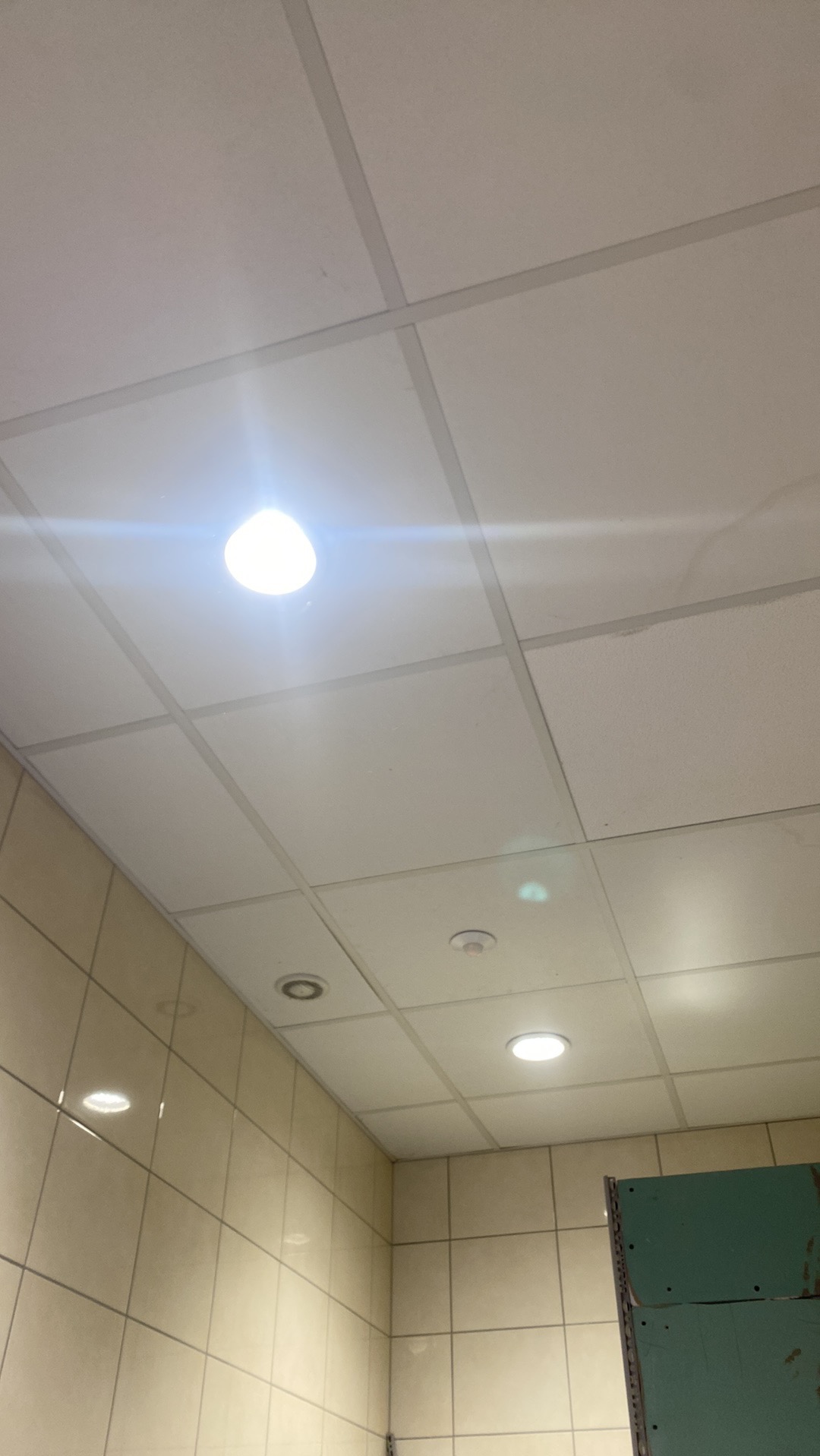 Kwadratowa podwieszana płyta sufitowa z zamontowanymi oprawami oświetleniowymi, w tym okrągły reflektor LED i czujnik dymu, z widocznymi kafelkami na ścianie.