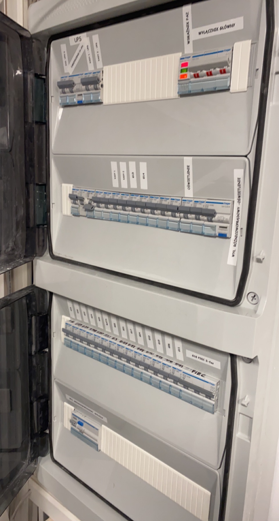 Otwarta skrzynka elektryczna z bezpiecznikami i naklejkami opisującymi obwody, widoczne wyłączniki główne, UPS i obwody oświetleniowe.