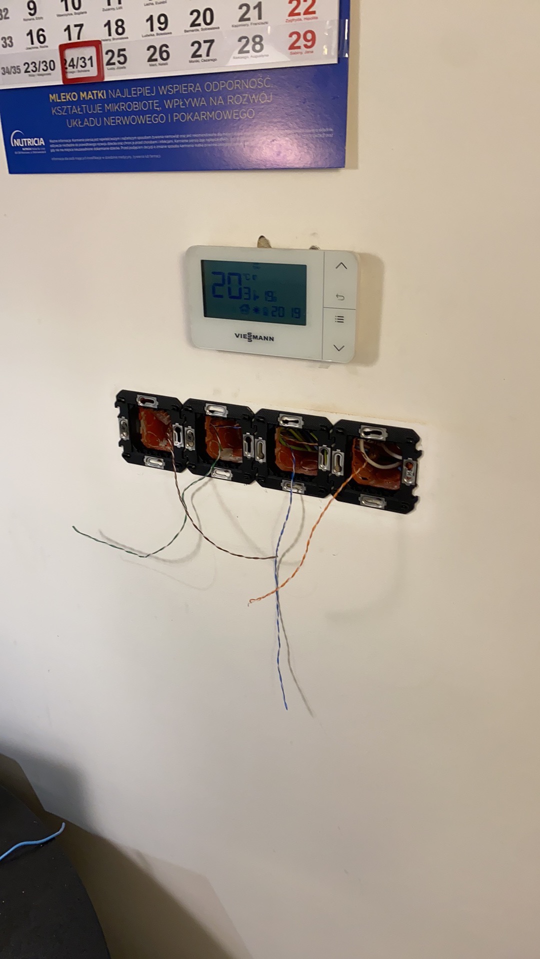 Cztery puszki elektryczne podtynkowe z wystającymi przewodami, zamontowane na białej ścianie pod termostatem Viessmann i kalendarzem Nutricia
