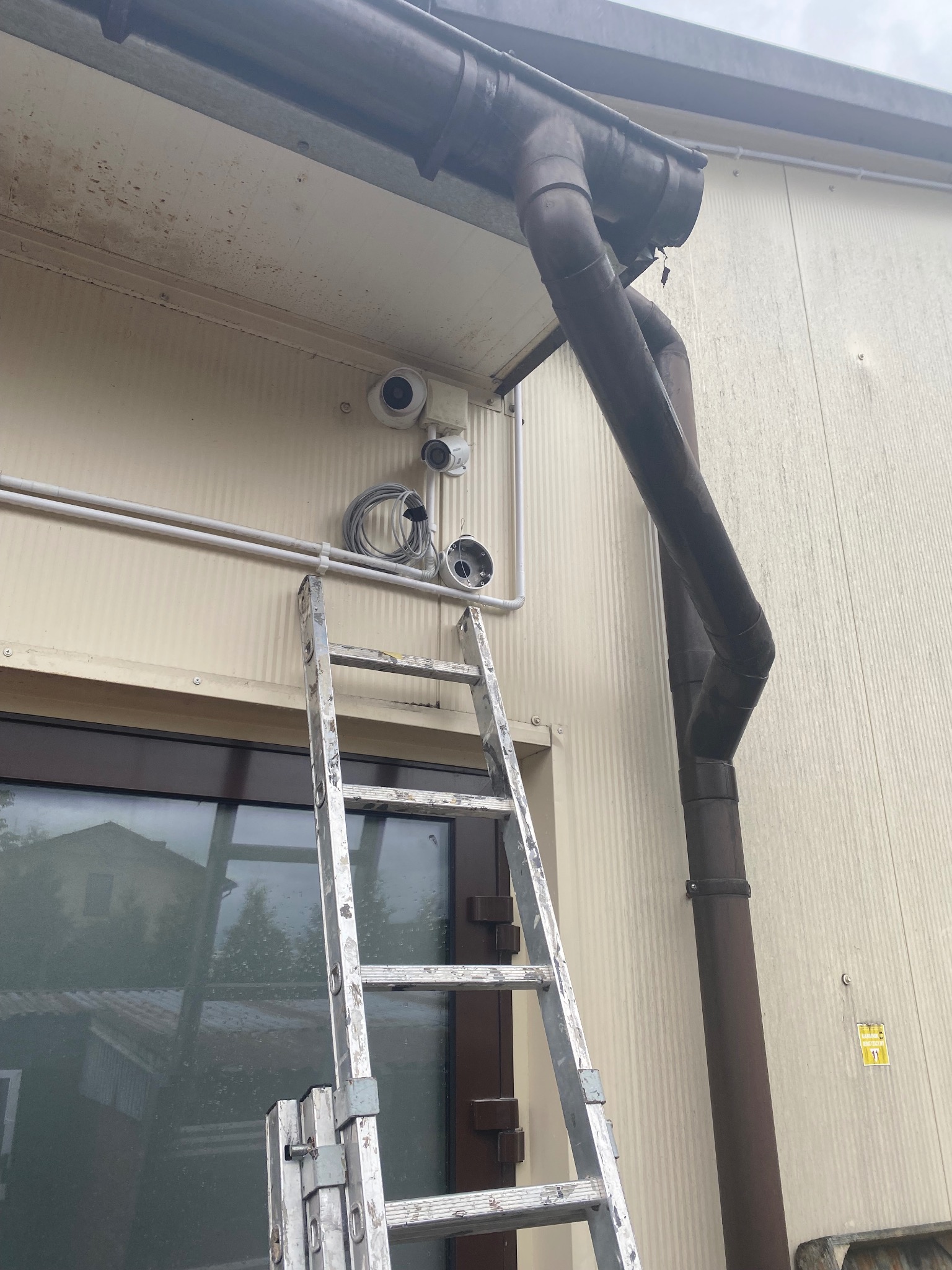 Montaż kamer monitoringu na elewacji budynku w Nawojowej, widoczna drabina oparta o ścianę, kamery w trakcie instalacji, przewody i rury prowadzone po ścianie.