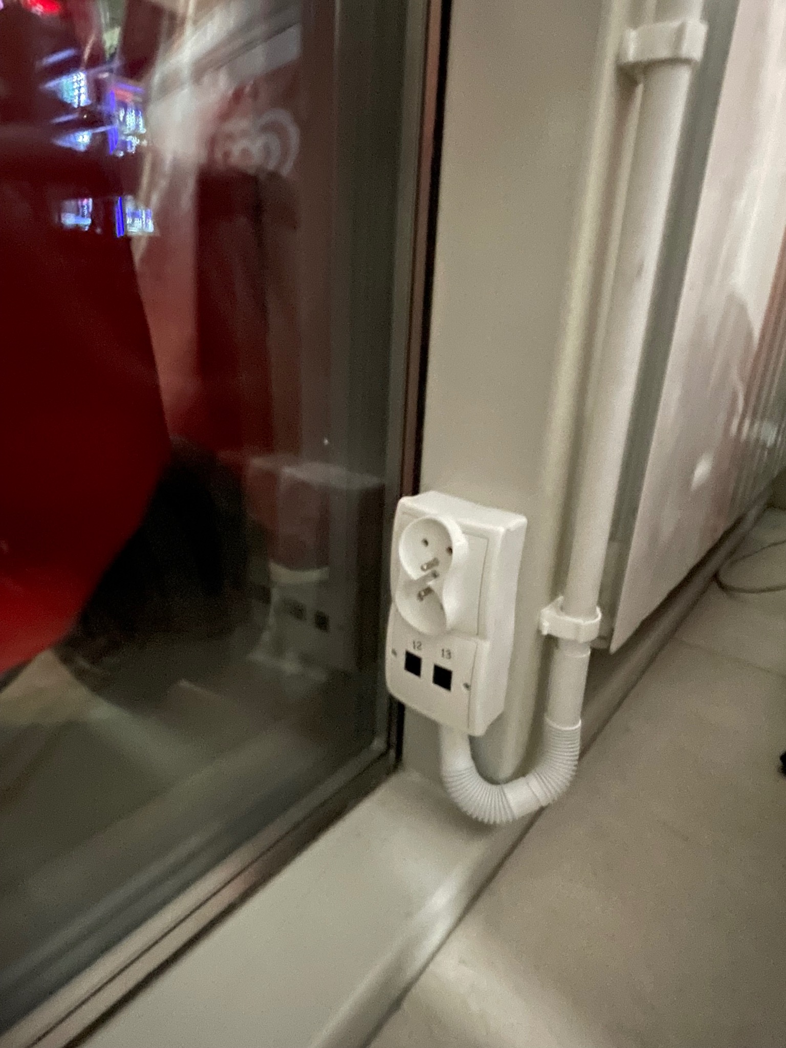 Białe gniazdko elektryczne podwójne z widocznymi stykami uziemiającymi, zamontowane na ścianie obok okna, z podłączonym przewodem w peszlu.