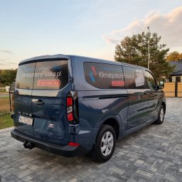 KLIMATYZATORY POLSKA - Niebieski samochód dostawczy Ford Custom z reklamą firmy od klimatyzacji na bocznych i tylnych szybach, zaparkowany na szarej kostce brukowej przed domem z drewnianym ogrodzeniem.