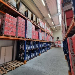 KLIMATYZATORY POLSKA - Magazyn z regałami wypełnionymi kartonami i elementami wentylacyjnymi, widok w perspektywie wzdłuż korytarza między regałami.