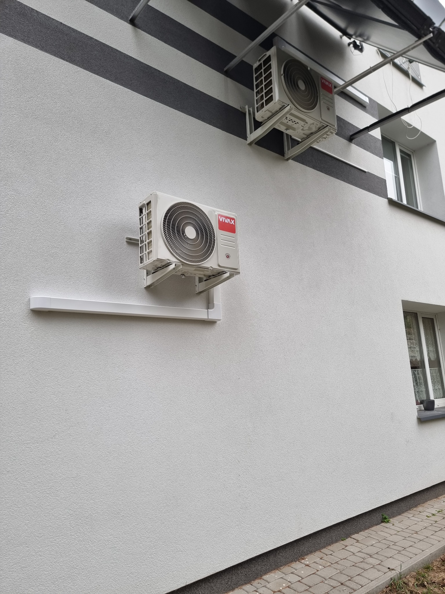 Dwa białe klimatyzatory marki Vivax zamontowane na jasnej elewacji budynku z szarymi pasami dekoracyjnymi i oknami.