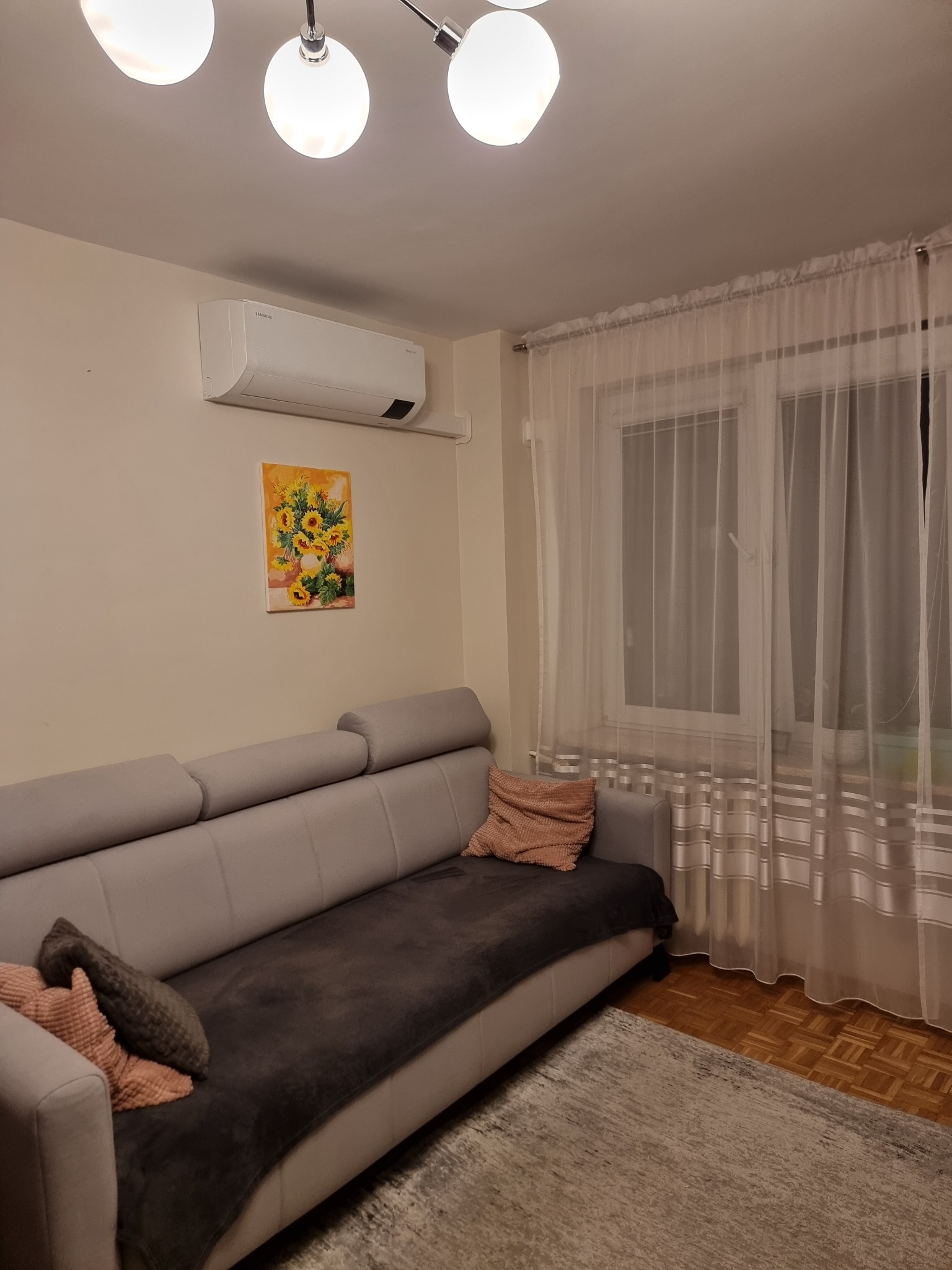 Biały klimatyzator Samsung zamontowany na ścianie nad szarym narożnikiem w salonie, pod nim obraz z motywem słoneczników, w tle okno z firankami.