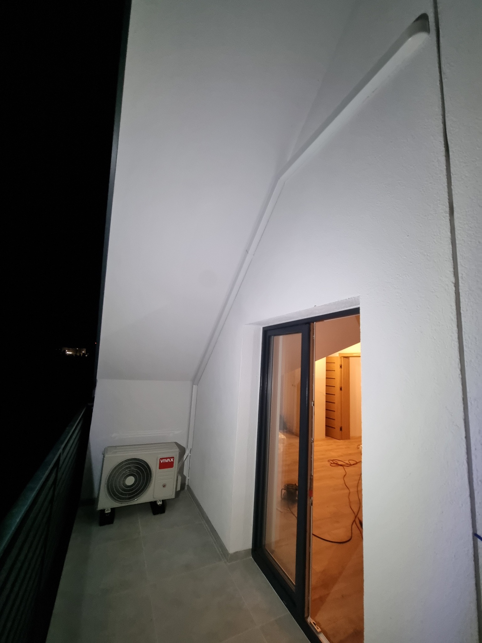Biała jednostka zewnętrzna klimatyzacji Vivax na balkonie z szarą podłogą, widok z dołu, z otwartymi drzwiami balkonowymi na jasne wnętrze.