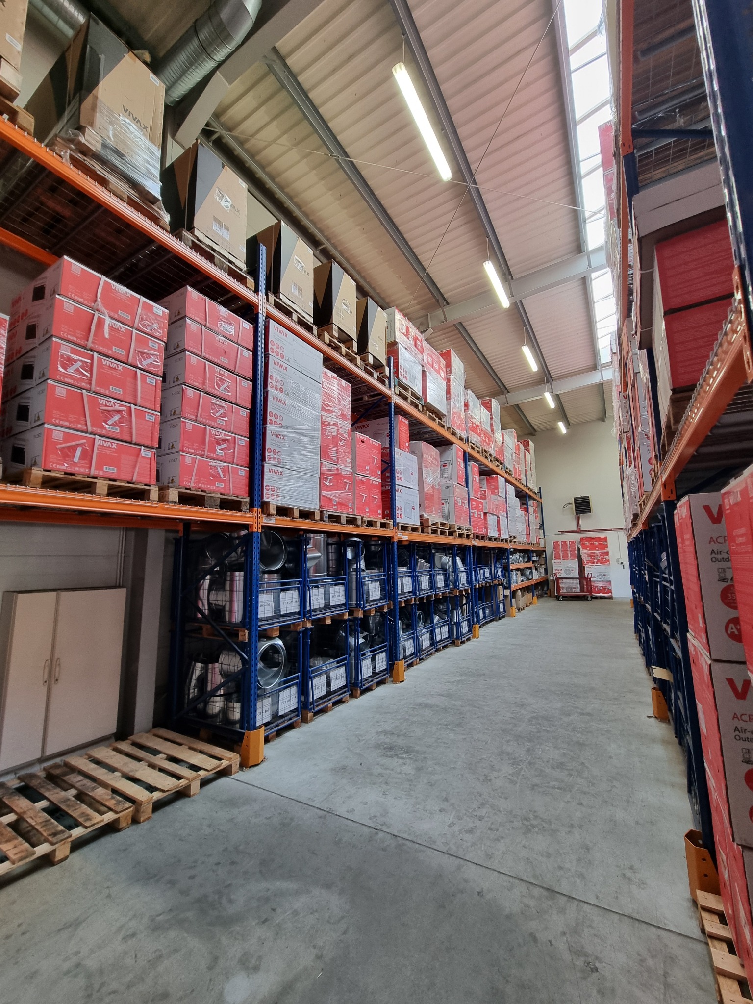 Magazyn z regałami wypełnionymi kartonami i elementami wentylacyjnymi, widok w perspektywie wzdłuż korytarza między regałami.