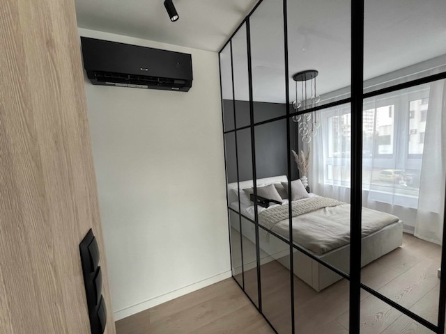 Czarna jednostka klimatyzacyjna na białej ścianie w sypialni z czarną, szklaną ścianką działową, widok na łóżko i okno.