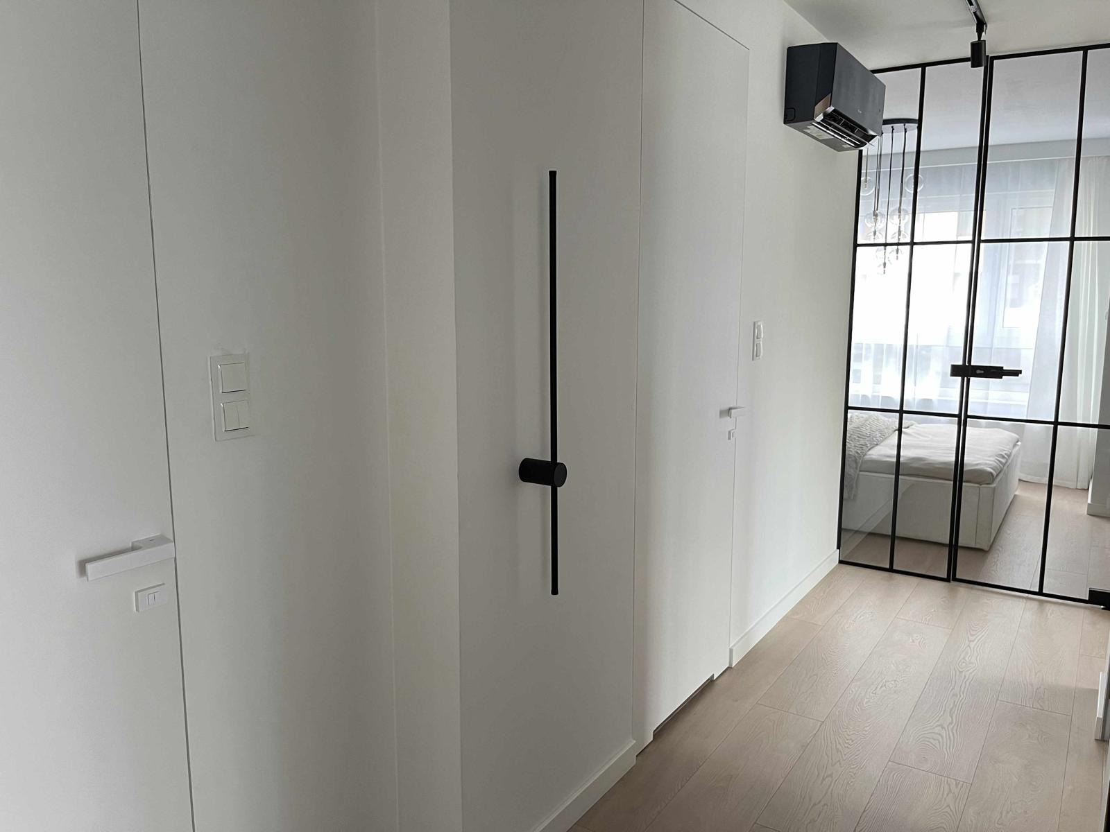 Czarny klimatyzator ścienny w minimalistycznym, białym wnętrzu z przeszklonymi drzwiami do sypialni.