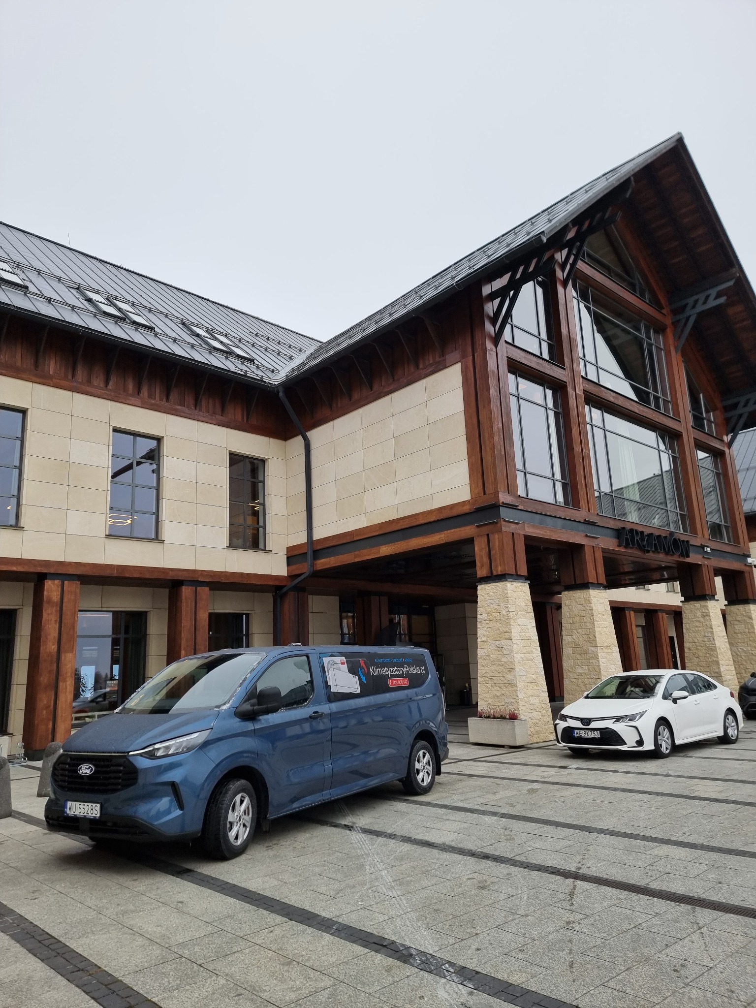 Niebieski samochód dostawczy z reklamą usług klimatyzacyjnych firmy z Lubartowa zaparkowany przed budynkiem z drewnianą konstrukcją i napisem 'TARNOW'.