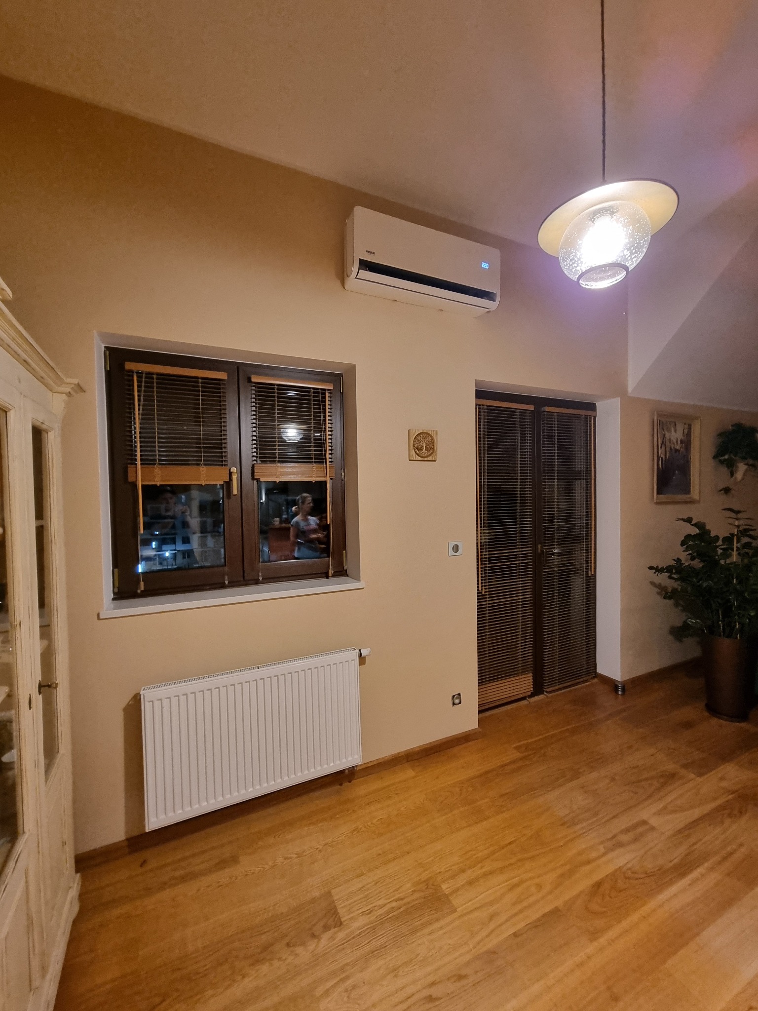 Biały klimatyzator ścienny w jasnym salonie z drewnianą podłogą i oknem z żaluzjami, oświetlony wiszącą lampą.