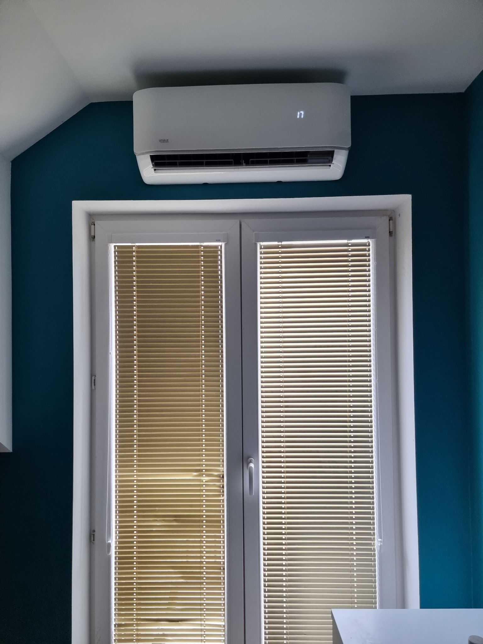 Biały klimatyzator na ścianie w pokoju z oknem z żaluzjami, wyświetlający temperaturę 17 stopni.