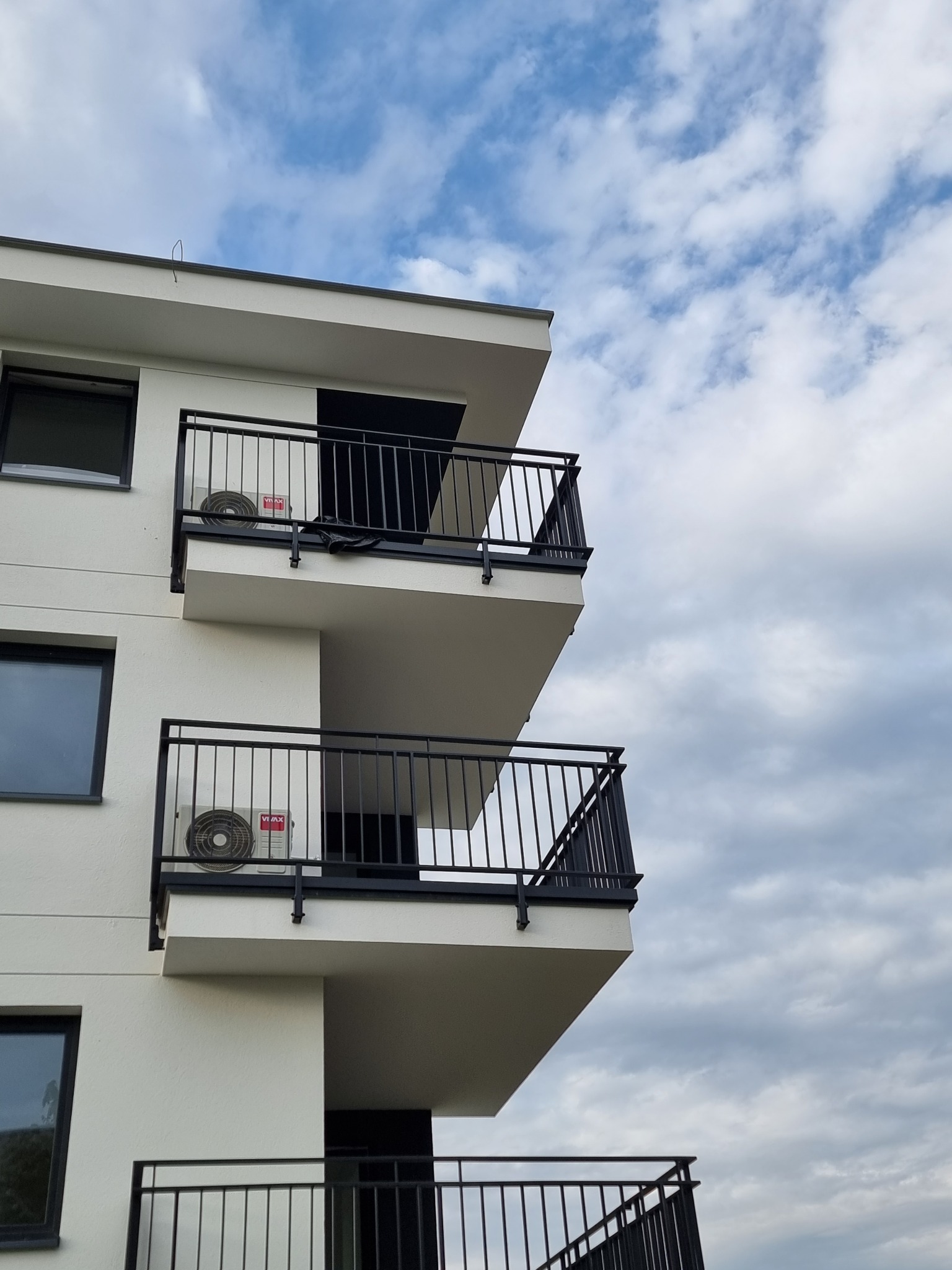 Biały blok mieszkalny z balkonami i zamontowanymi klimatyzatorami Vimax na każdym balkonie, na tle pochmurnego nieba.