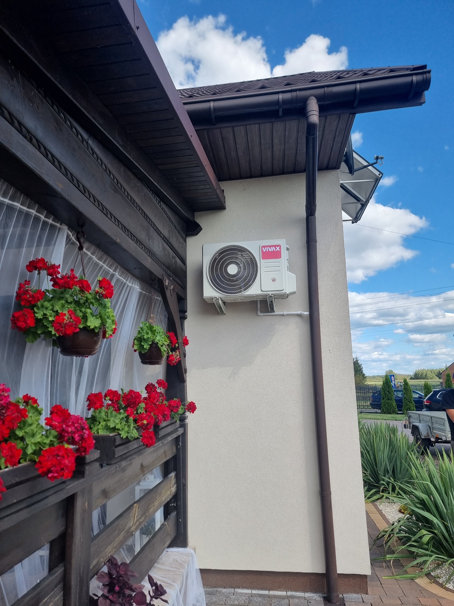 Biała jednostka zewnętrzna klimatyzacji VIVAX zamontowana na elewacji domu z brązowym systemem rynnowym, obok drewnianej werandy ozdobionej czerwonymi pelargoniami w wiszących donicach.