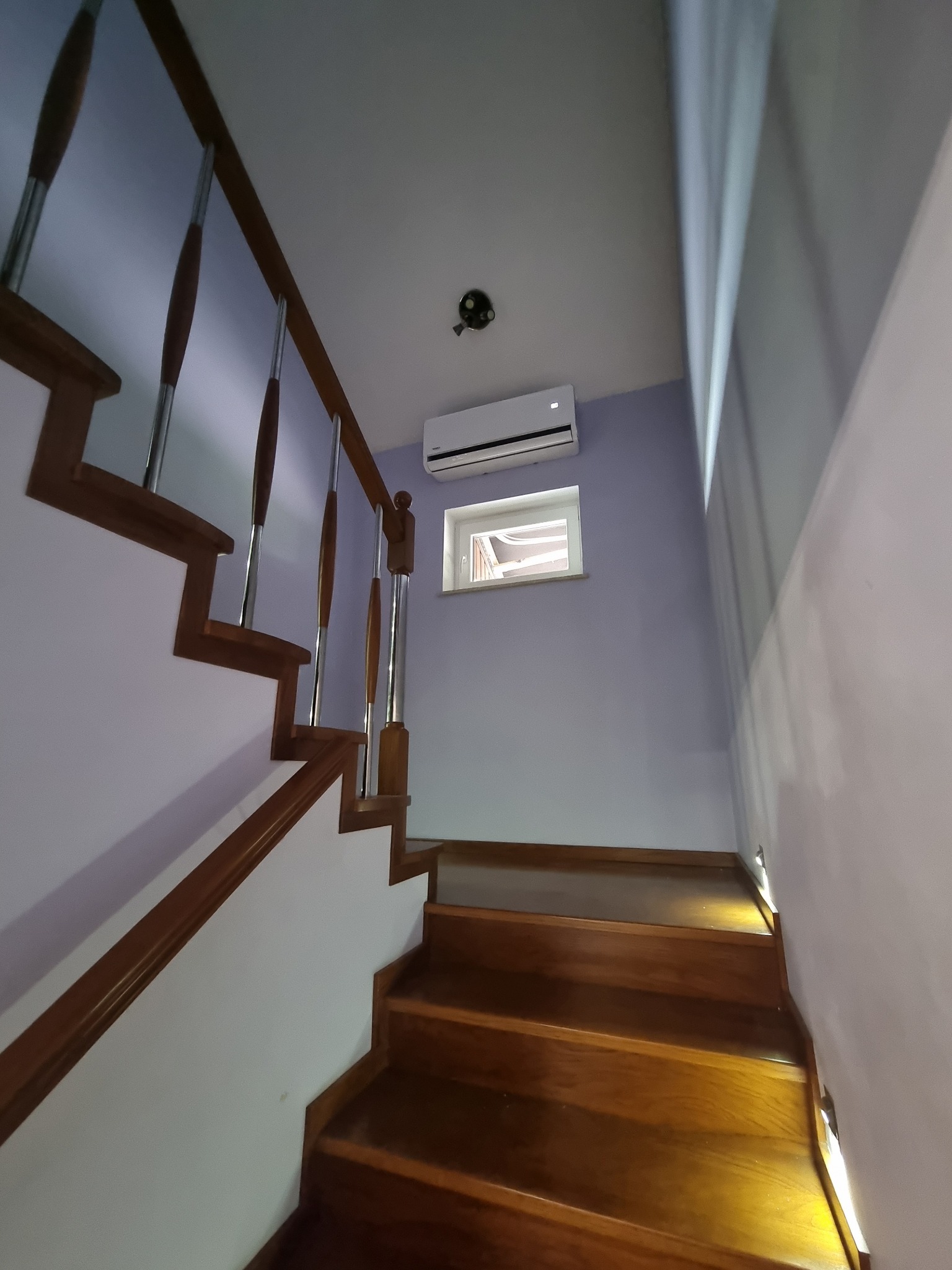 Biały klimatyzator ścienny zamontowany nad oknem na klatce schodowej z drewnianymi schodami i metalowymi poręczami.