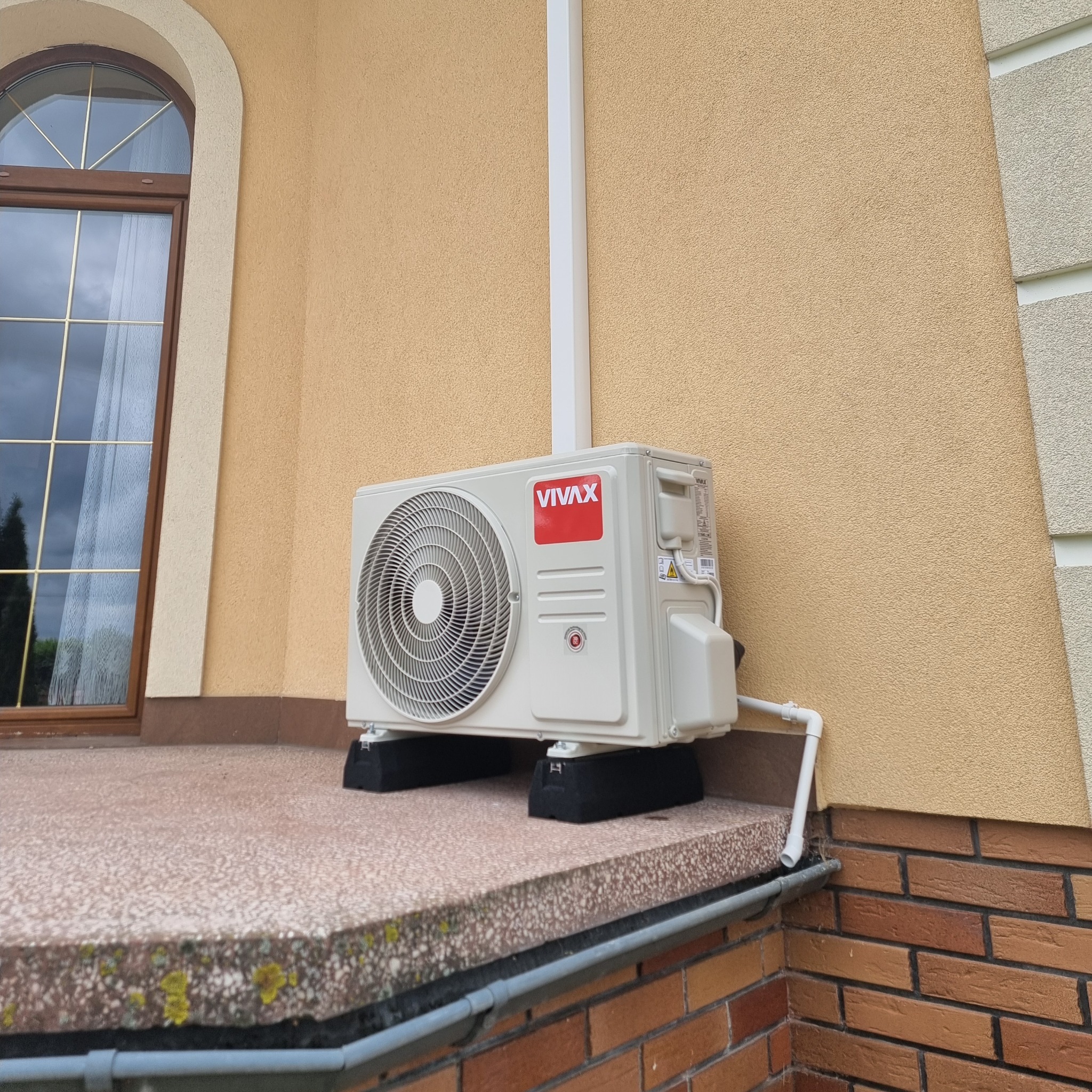Zewnętrzna jednostka klimatyzacji marki Vivax zamontowana na wspornikach na tarasie domu z beżową elewacją i brązową cegłą.