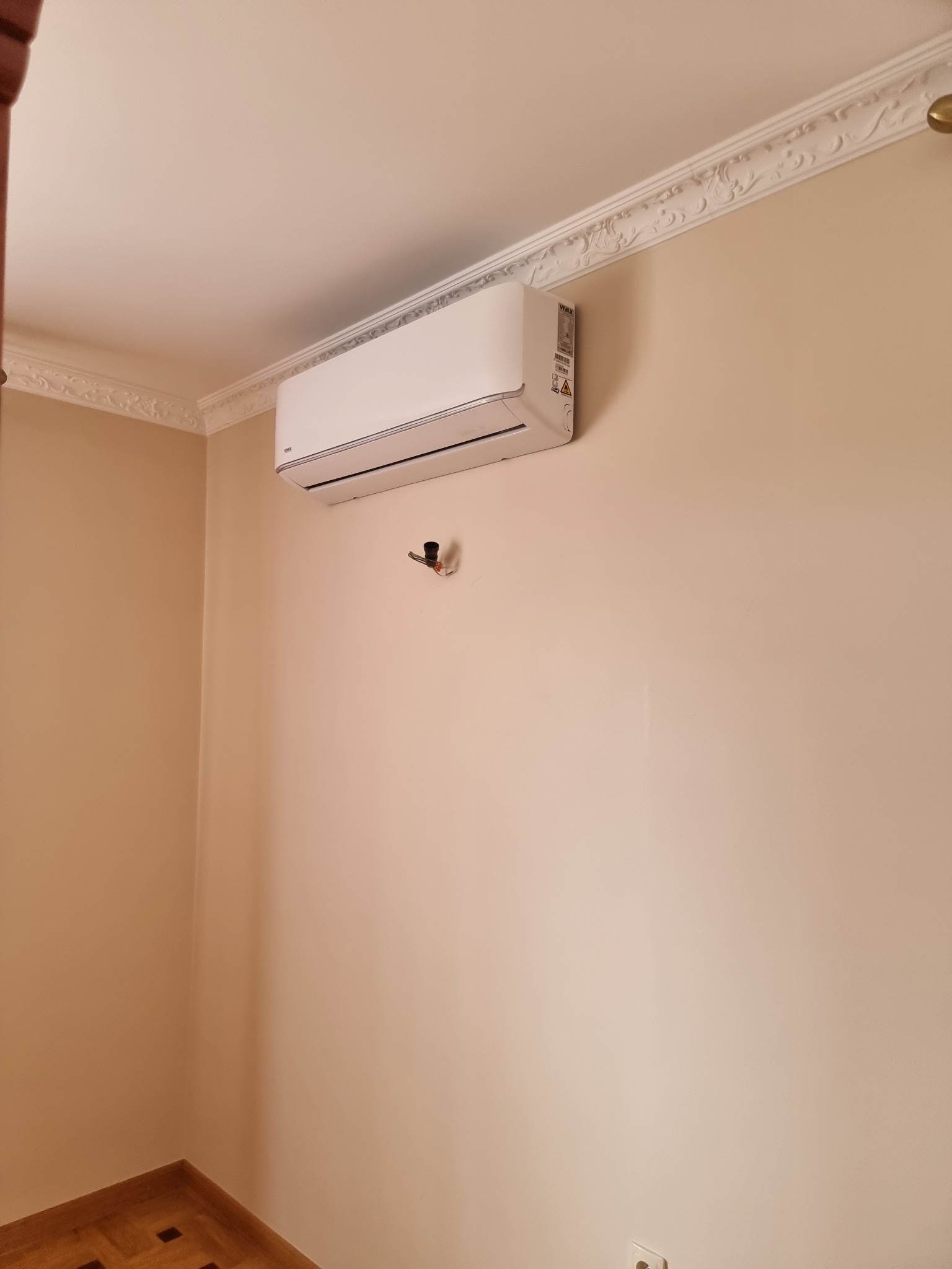 Biała klimatyzacja ścienna zamontowana w pokoju z dekoracyjną listwą sufitową i parkietem.