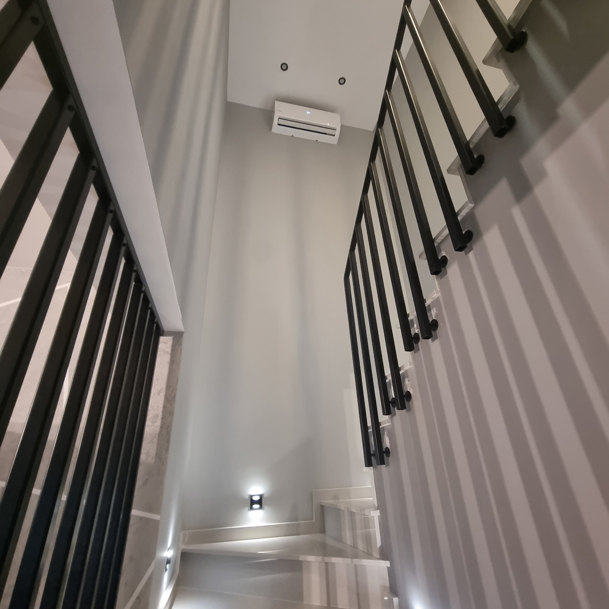 Wnętrze domu z klimatyzatorem zamontowanym pod sufitem, widoczne schody z czarną balustradą i oświetleniem punktowym.