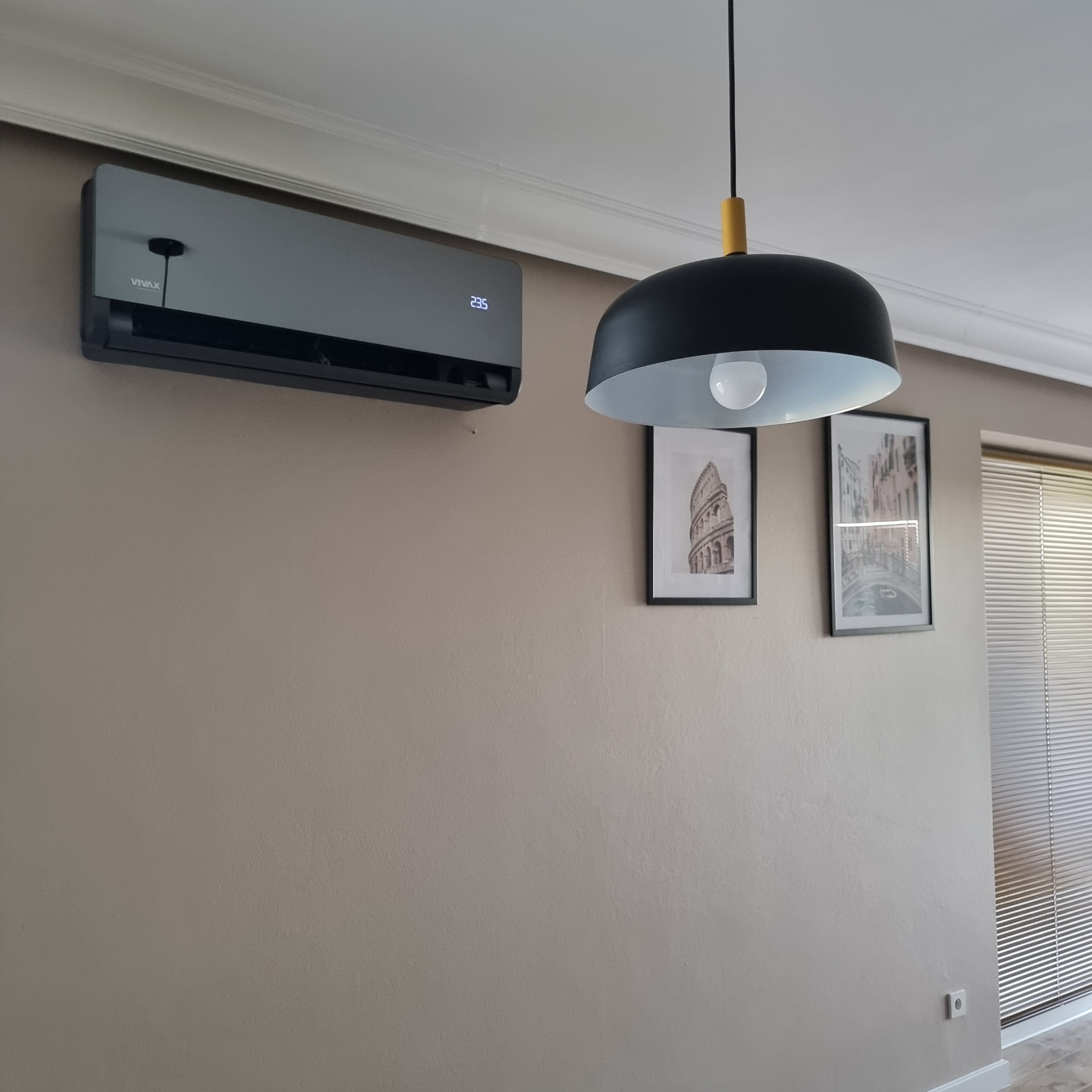 Szary klimatyzator Vivax zamontowany na ścianie w pomieszczeniu mieszkalnym, z widocznym wyświetlaczem temperatury, czarna lampa wisząca z elementami drewna, obrazy z motywami architektonicznymi...