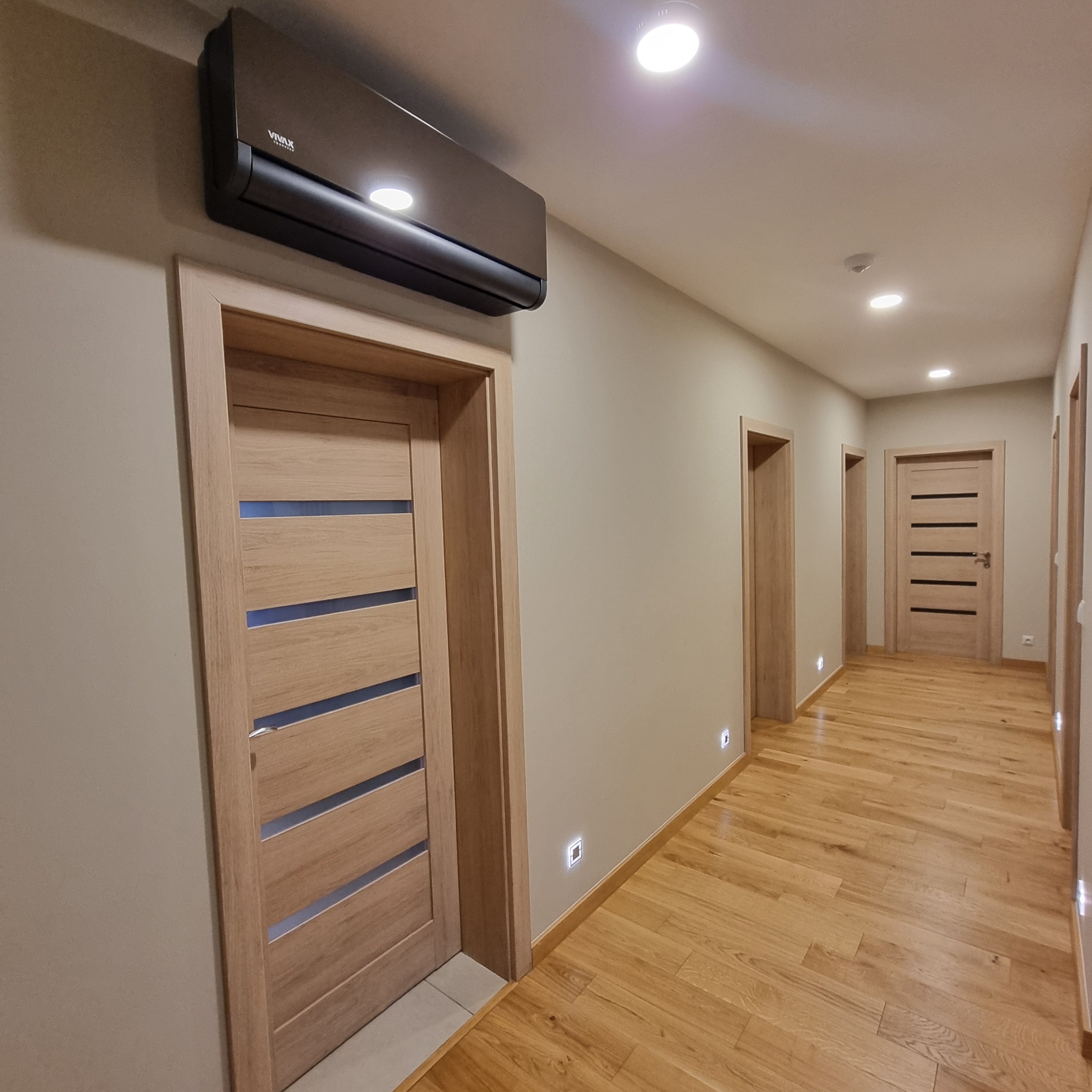 Ciemna jednostka klimatyzacyjna VIVAX zamontowana nad drewnianymi drzwiami w jasnym korytarzu z drewnianą podłogą i punktowymi lampkami ściennymi.