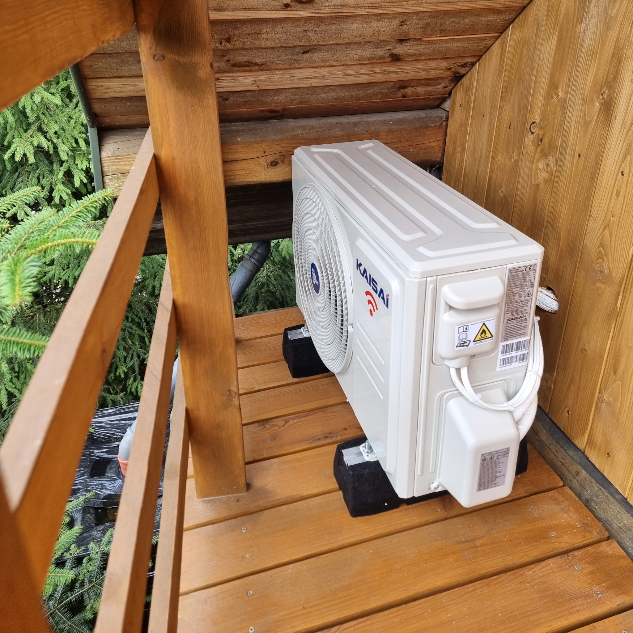Zewnętrzna jednostka klimatyzatora Kaisai zamontowana na drewnianym balkonie, widok z góry, czarne podkładki antywibracyjne pod urządzeniem.
