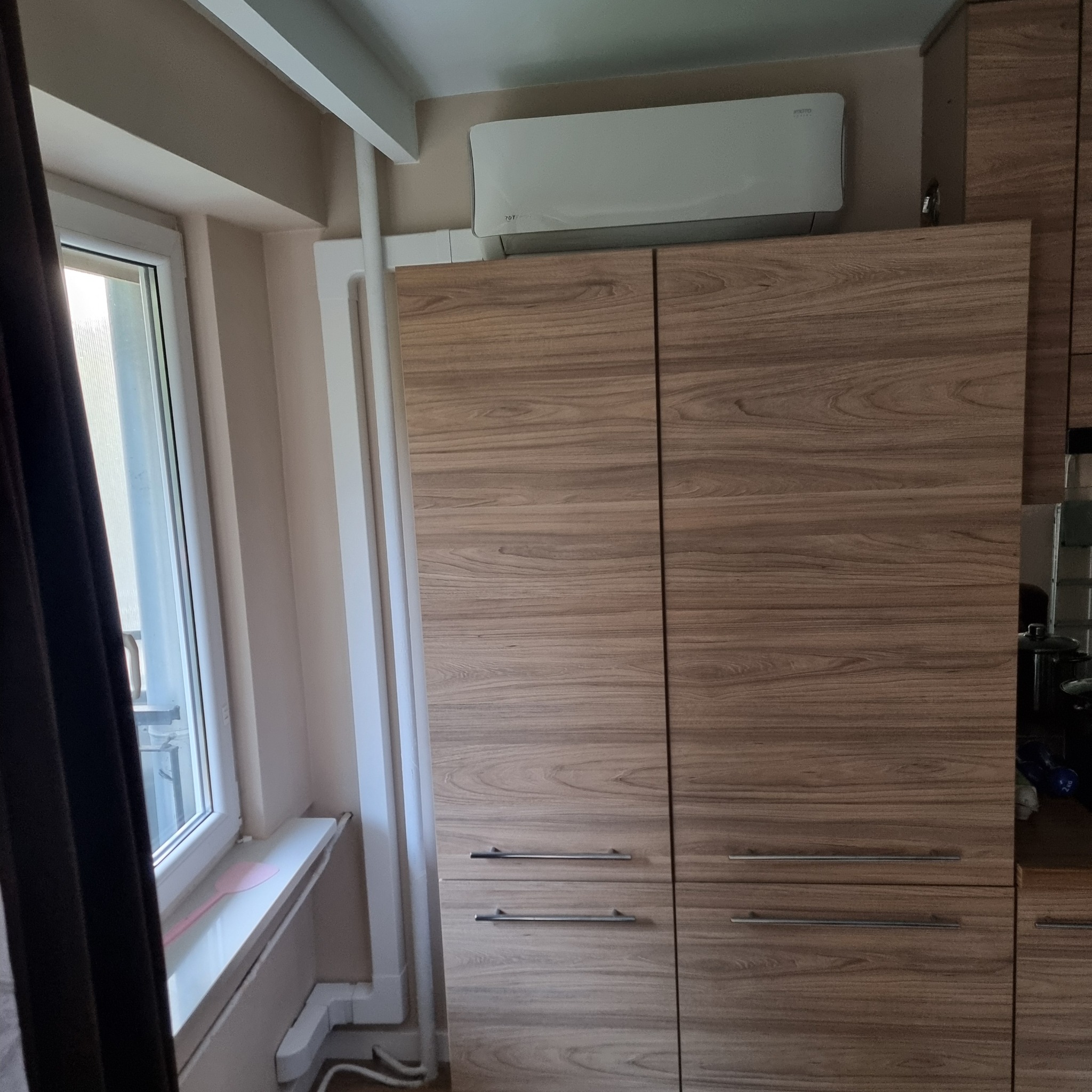 Biała jednostka klimatyzacyjna zamontowana nad drewnianą szafą w pokoju obok okna.
