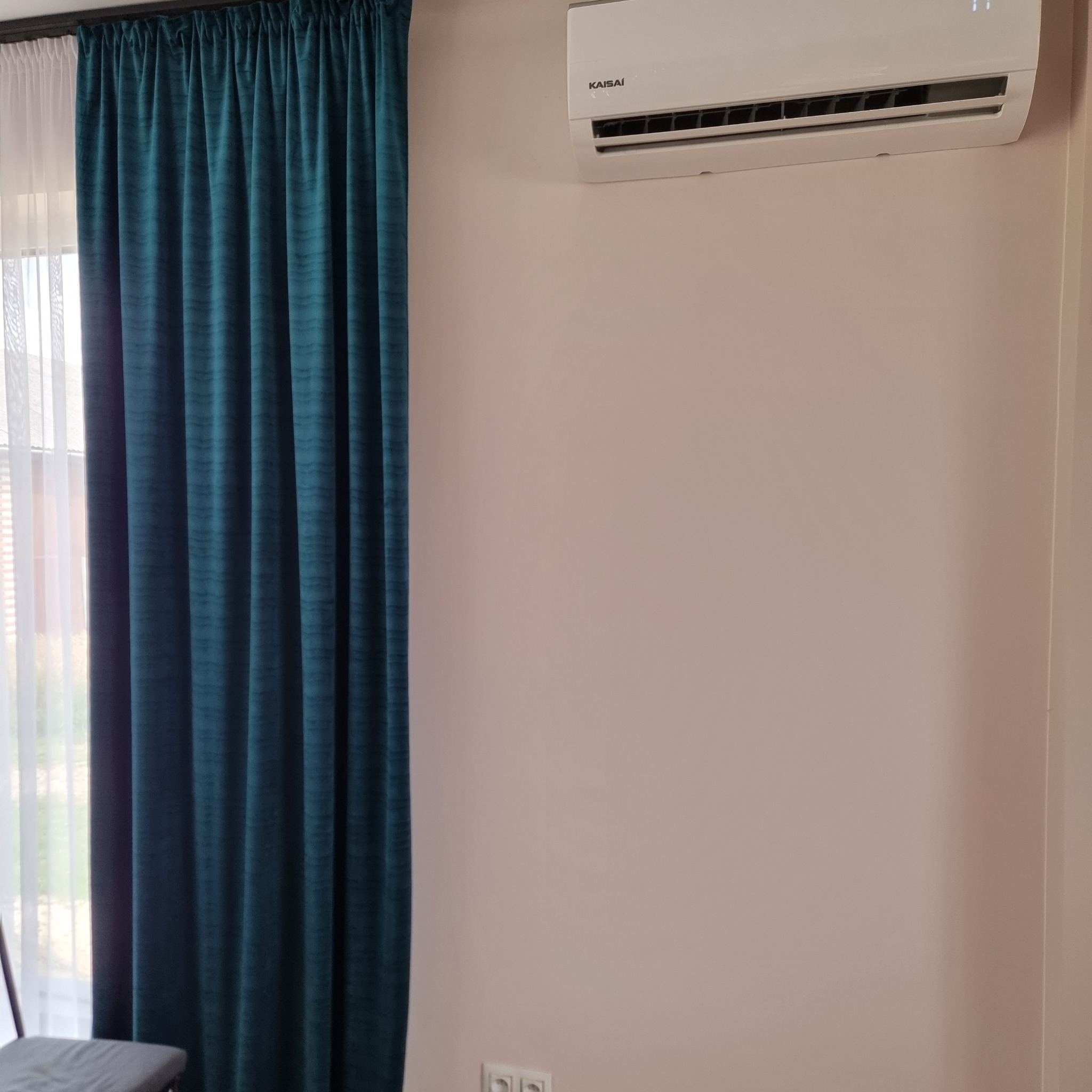 Zamontowany klimatyzator Kaisaí na ścianie w pokoju z zasłoniętym oknem, widoczne gniazdka elektryczne.