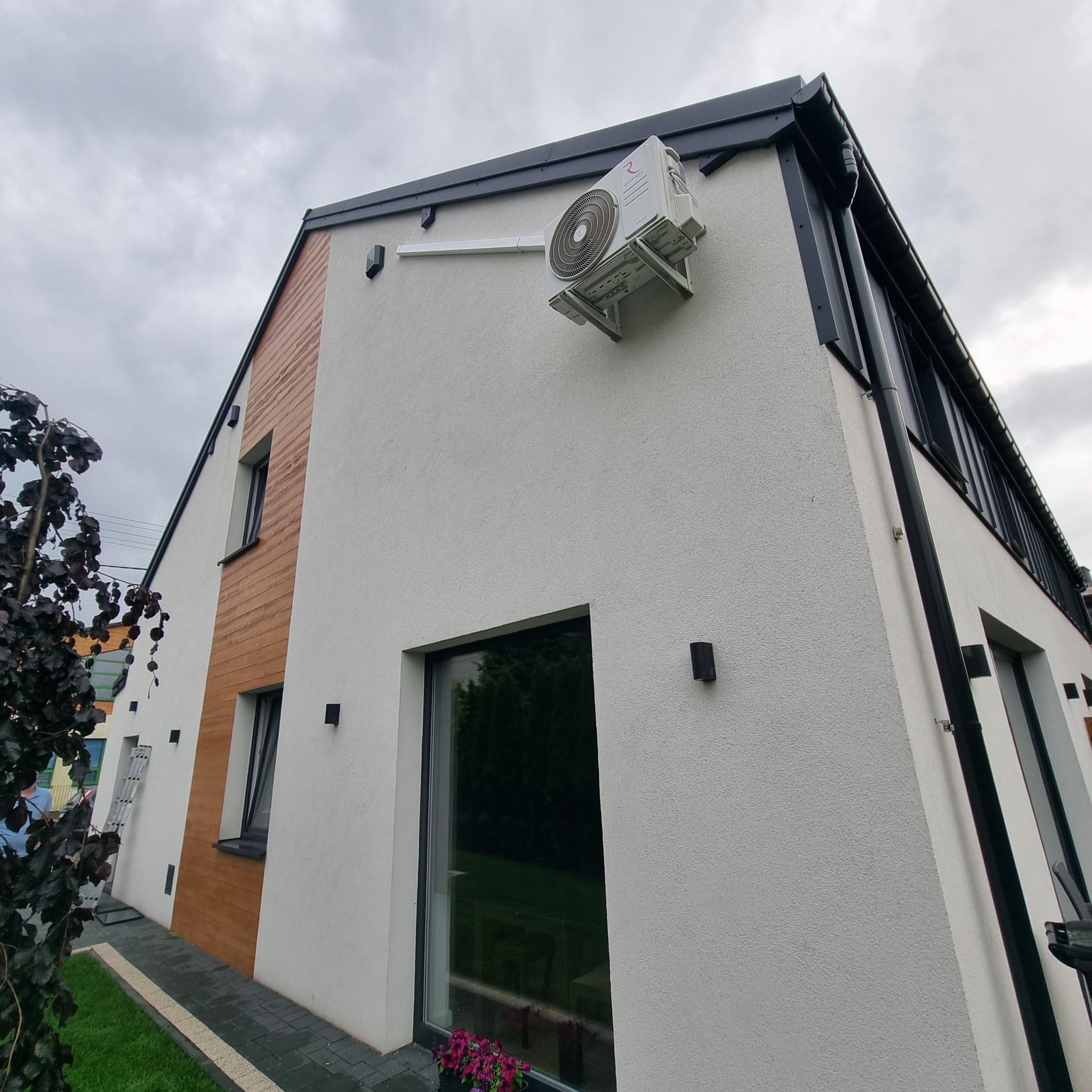 Zewnętrzna jednostka klimatyzatora zamontowana na ścianie nowoczesnego domu z drewnianą elewacją, widok z dołu.