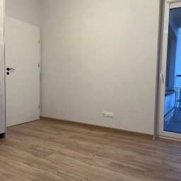 FixPoint - Pusta, jasna przestrzeń po remoncie: drewnopodobne panele podłogowe, białe ściany z gniazdkami i białe drzwi z czarną klamką, balkon z widokiem na zewnątrz.